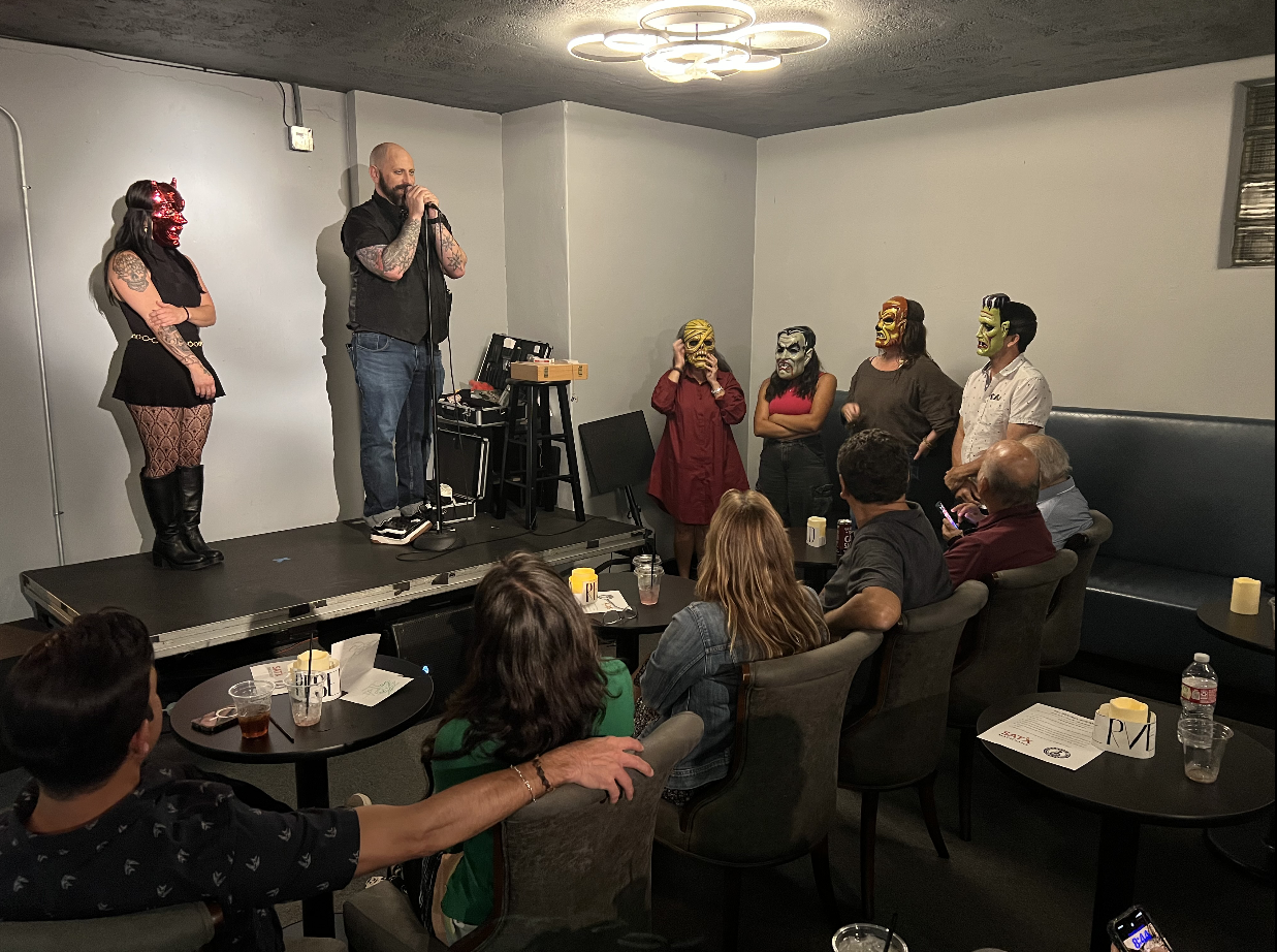 Mask performers performing on stage at a comedy or entertainment show, with audience seated at tables watching.
