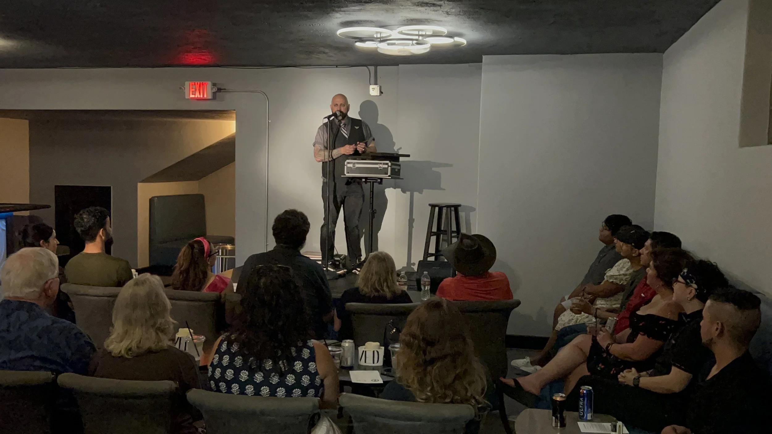 A man performs stand-up comedy on stage at a small venue with an audience seated around tables, some with drinks. The stage has a microphone, a speaker, and a stool, with lighting fixtures above.