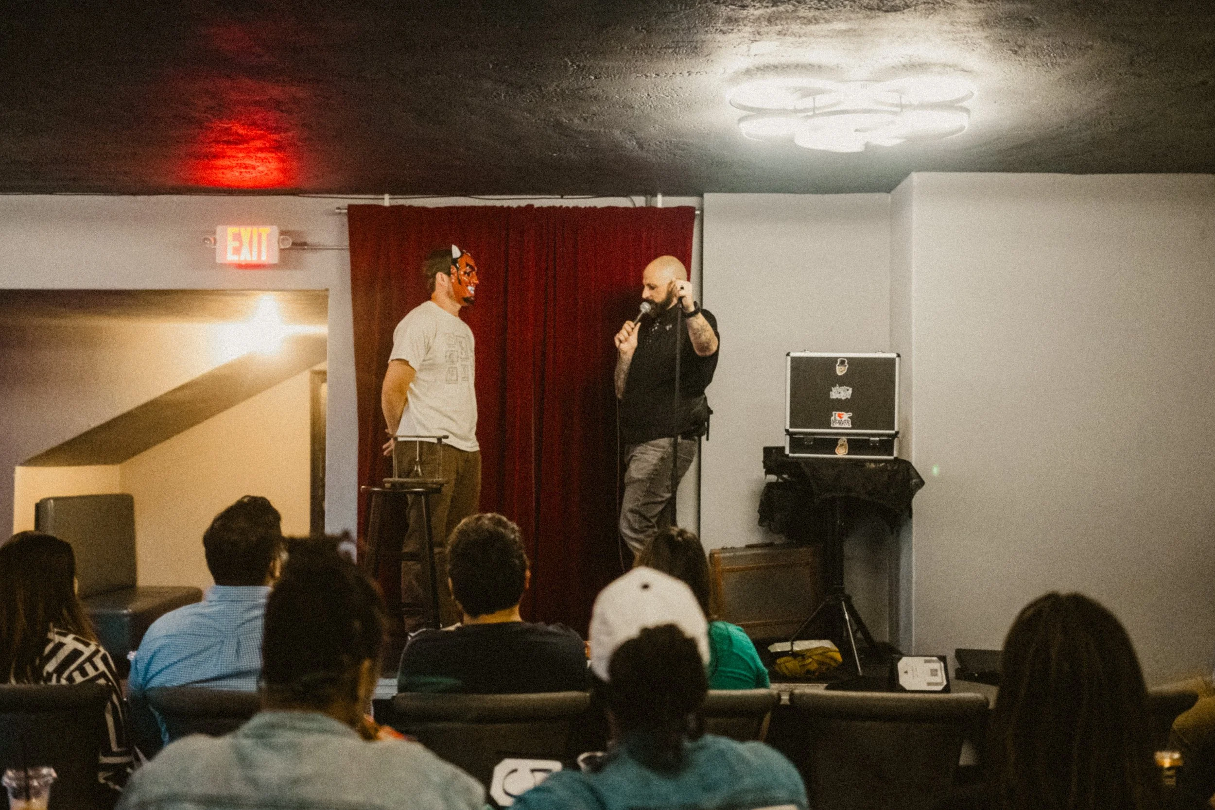 A comedy show with two performers on stage, one wearing a tiger mask and the other holding a microphone, in front of an audience seated in a dark room with a red curtain background.