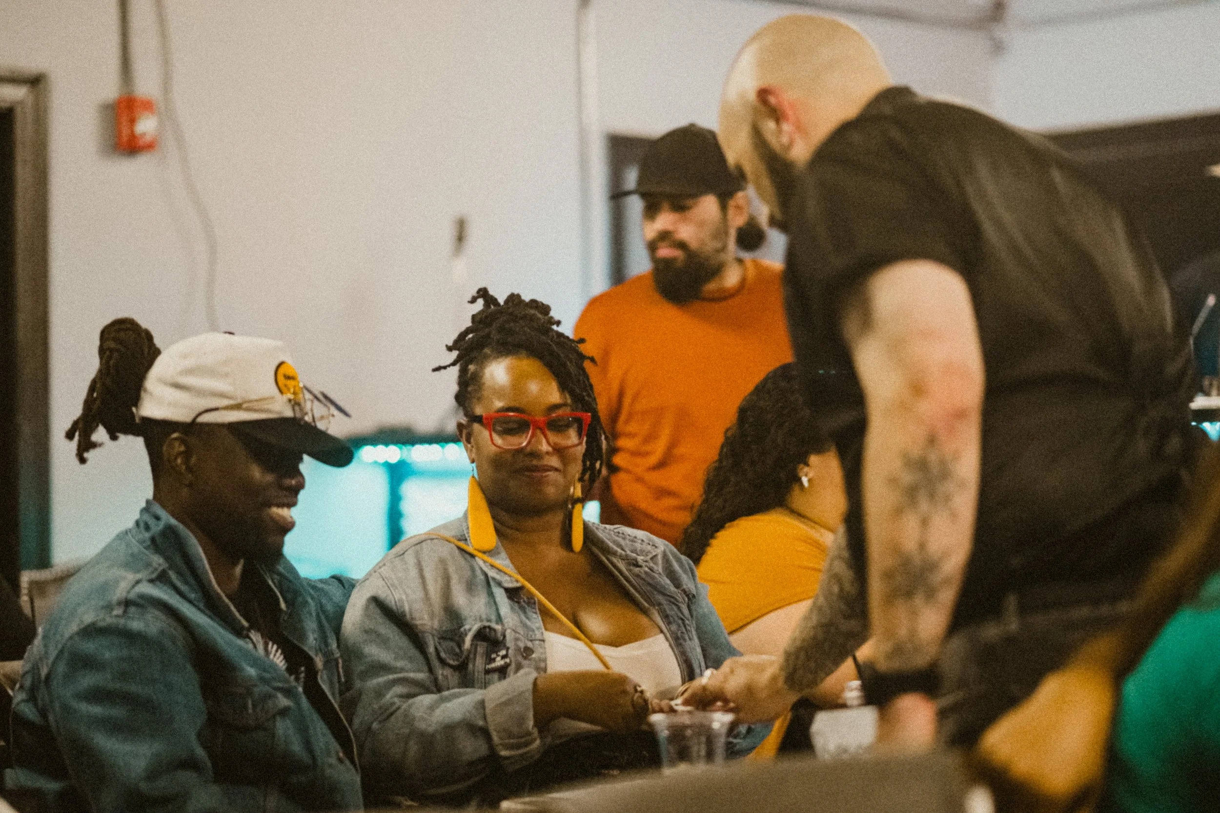 Group of people socializing indoors, some wearing colorful clothing and accessories, with a mix of seated and standing individuals.