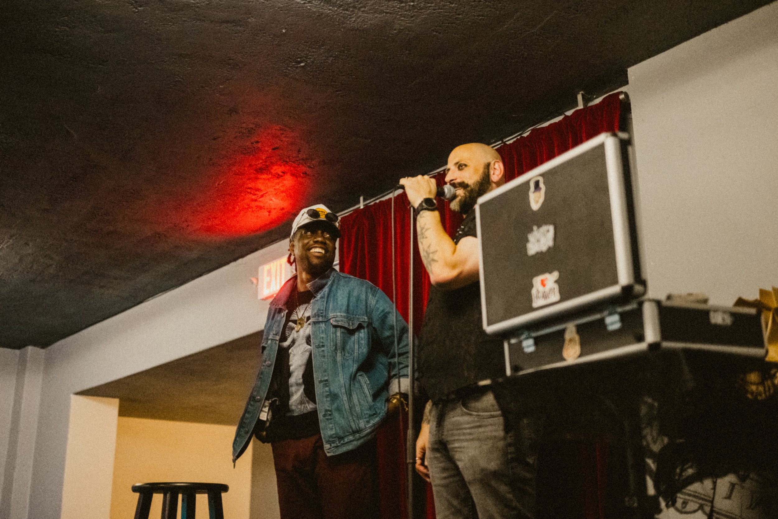 Two men stand on a stage inside a venue with a red curtain backdrop. A magician is speaking into a microphone, and the other is smiling and looking at him. There is a black case with stickers on it and a stool nearby.