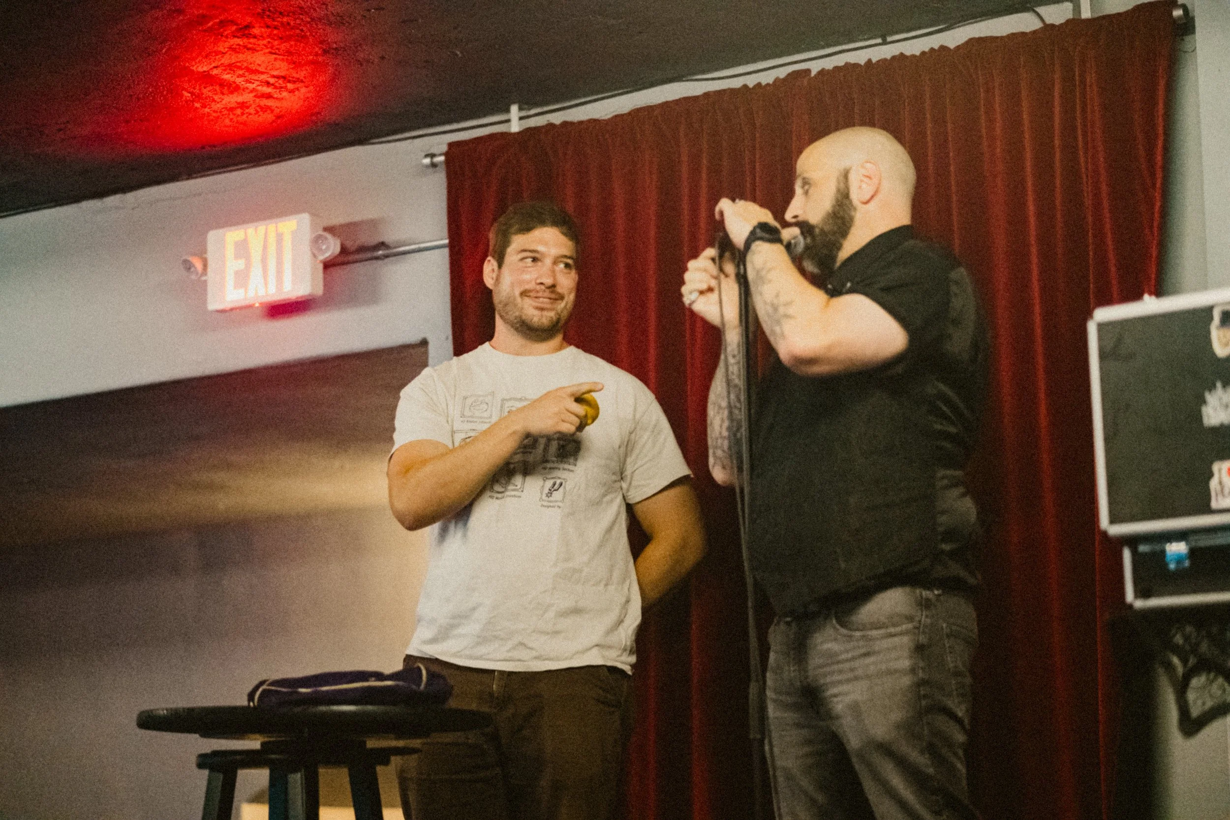 Two men on stage, one holding a microphone and speaking, the other smiling and pointing at him. Red curtains in the background and an exit sign above.