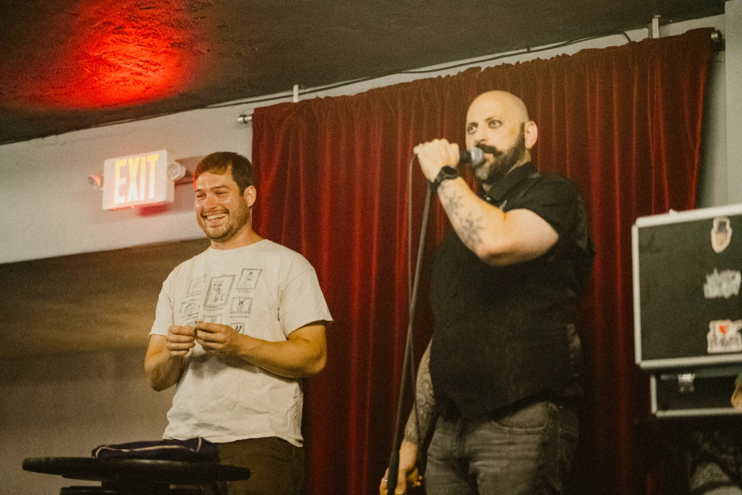 Two men on a stage with red curtains, one holding a microphone and the other smiling and holding a small object. There is an illuminated exit sign above them.