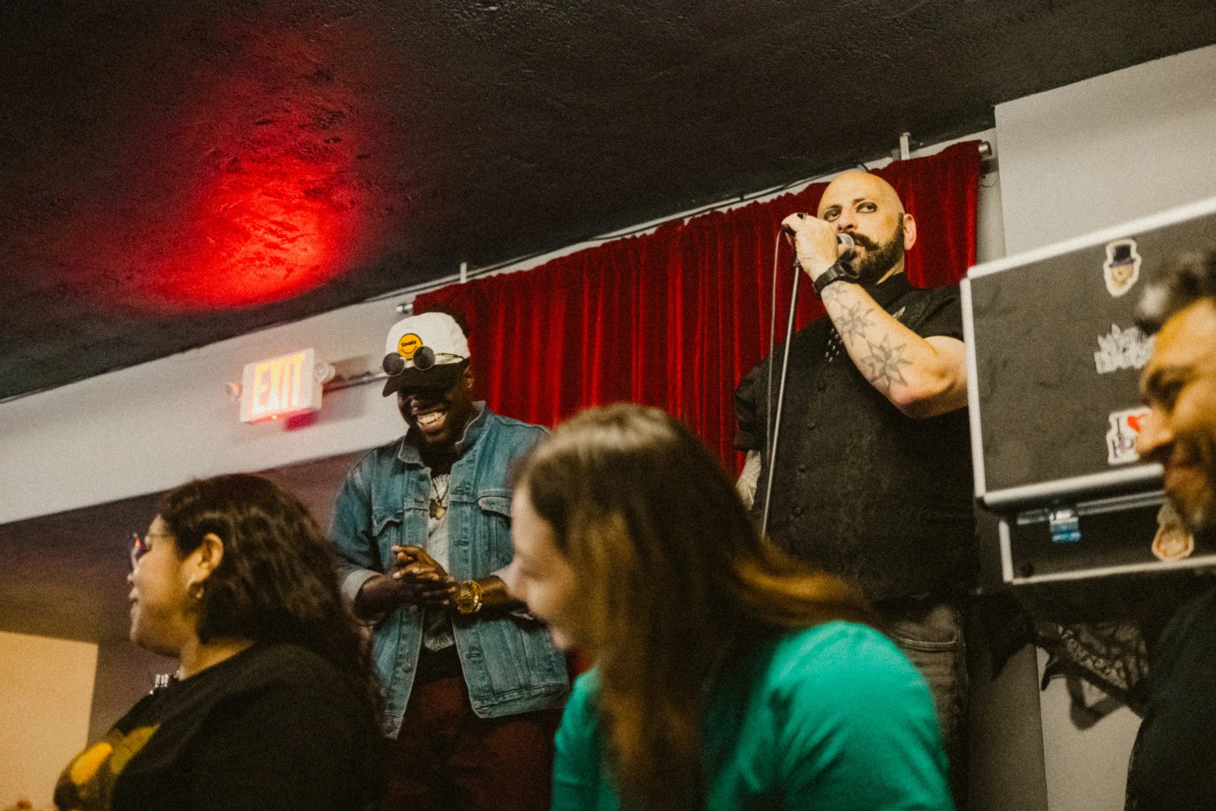 Two men are on stage with red curtain background, one is singing into a microphone and the other is smiling while looking at his phone. Several women are in the foreground.