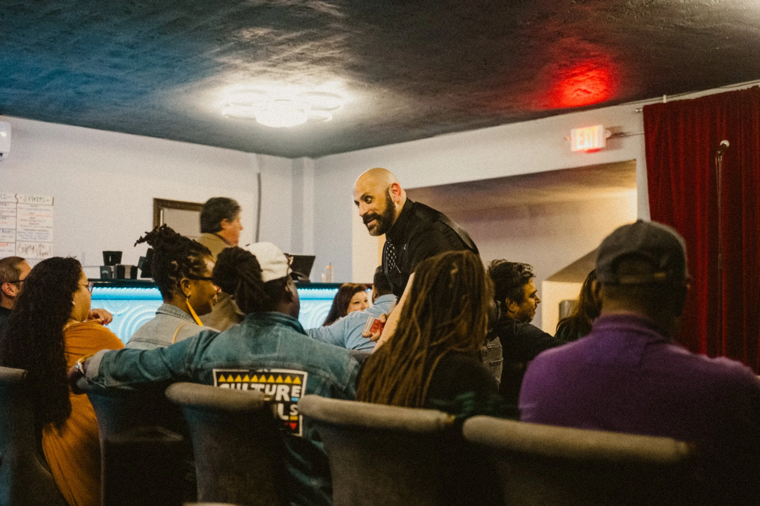 A man with a beard and a black shirt is speaking to a group of diverse people in a dimly lit room with a nightclub or bar setting. The audience includes men and women sitting at tables, and some are reaching out or engaging with the speaker.