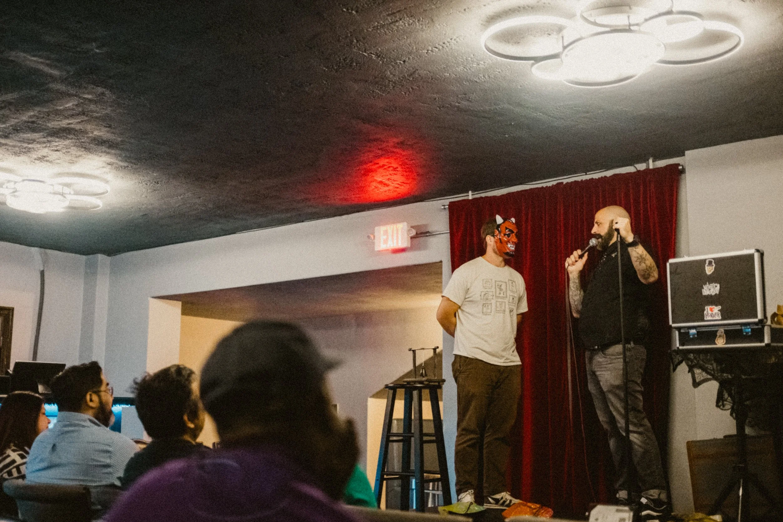 A comedy magic show on stage with audience member wearing a mask and casual clothes, and tTwo performers on a stage with a red curtain; one is speaking into a microphone, the other wears a red fox mask. Audience members are seated in front, watching.