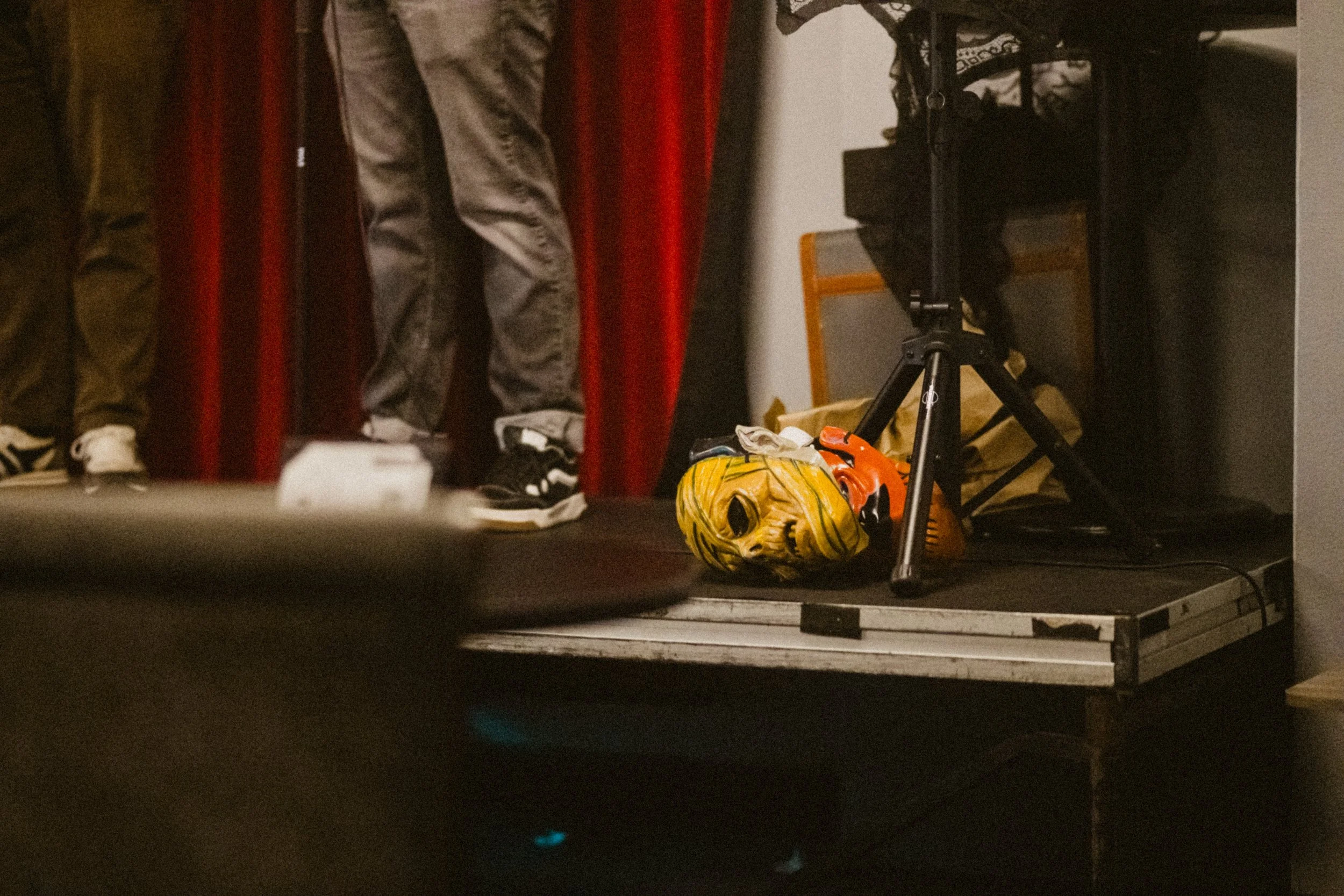 A Halloween mask with a painted face and flowing black hair lies on the floor next to a small orange toy chainsaw, with photography equipment and people standing in the background.