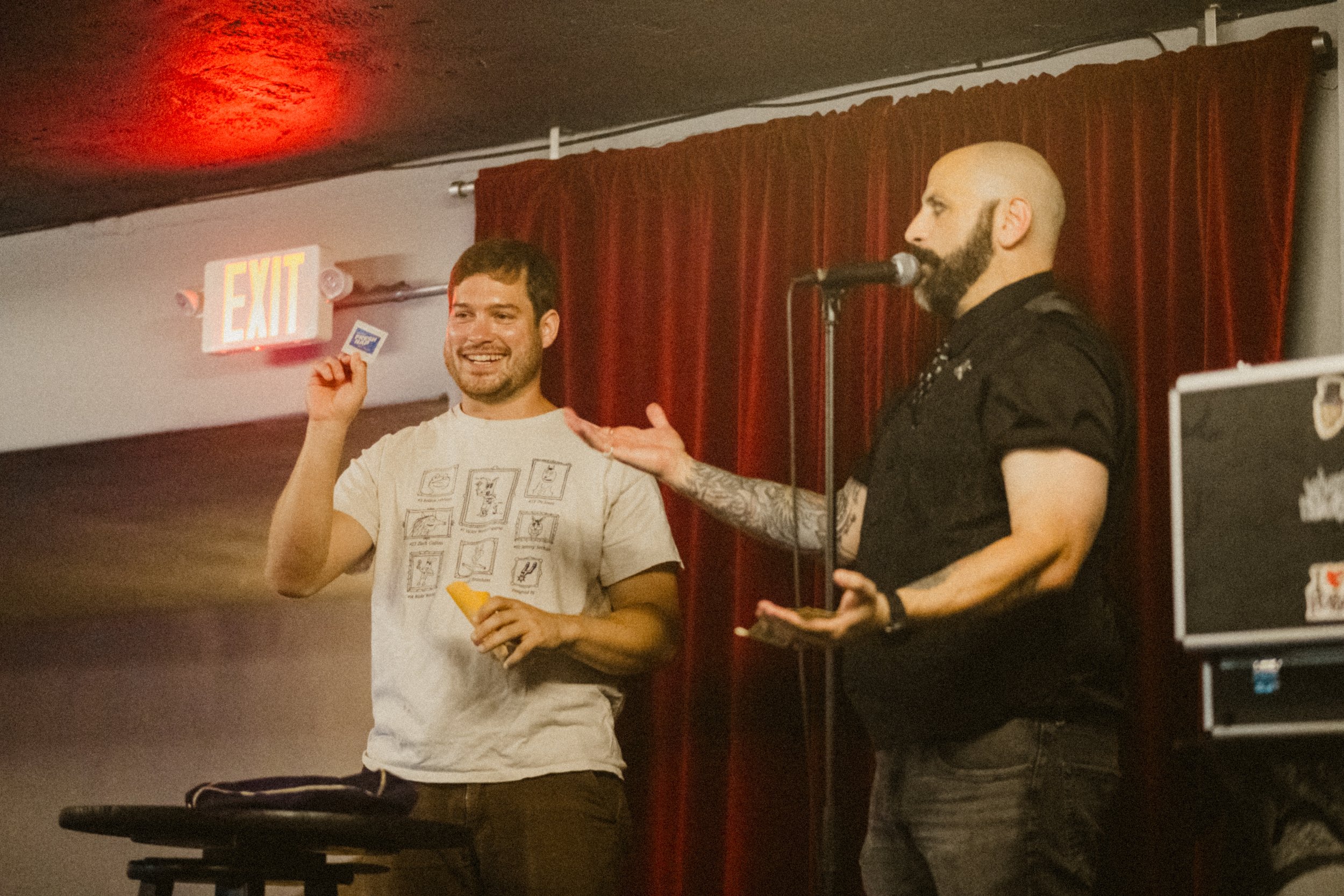 Two men on stage, one holding a card and smiling, the other speaking into a microphone, with a red curtain in the background.