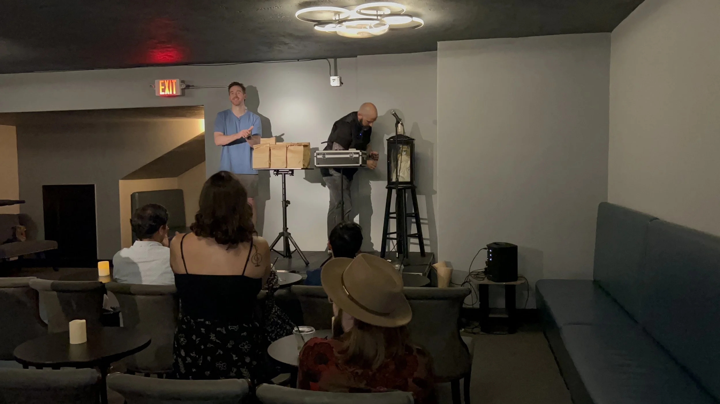 Two performers on a small stage, one man with a microphone and another man adjusting equipment, in a dimly lit room with an audience seated and watching.
