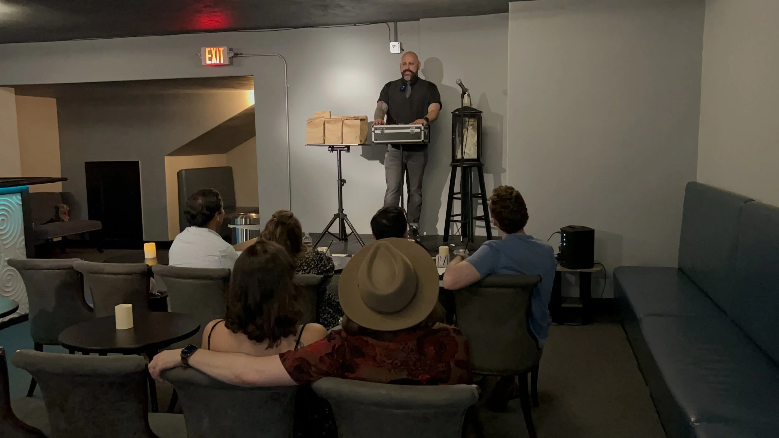 A man with a beard, wearing a black shirt, performs a magic trick on stage in front of an audience in a dimly lit room. The audience consists of five people seated at tables, watching him. The man is standing behind a small stand with objects and has