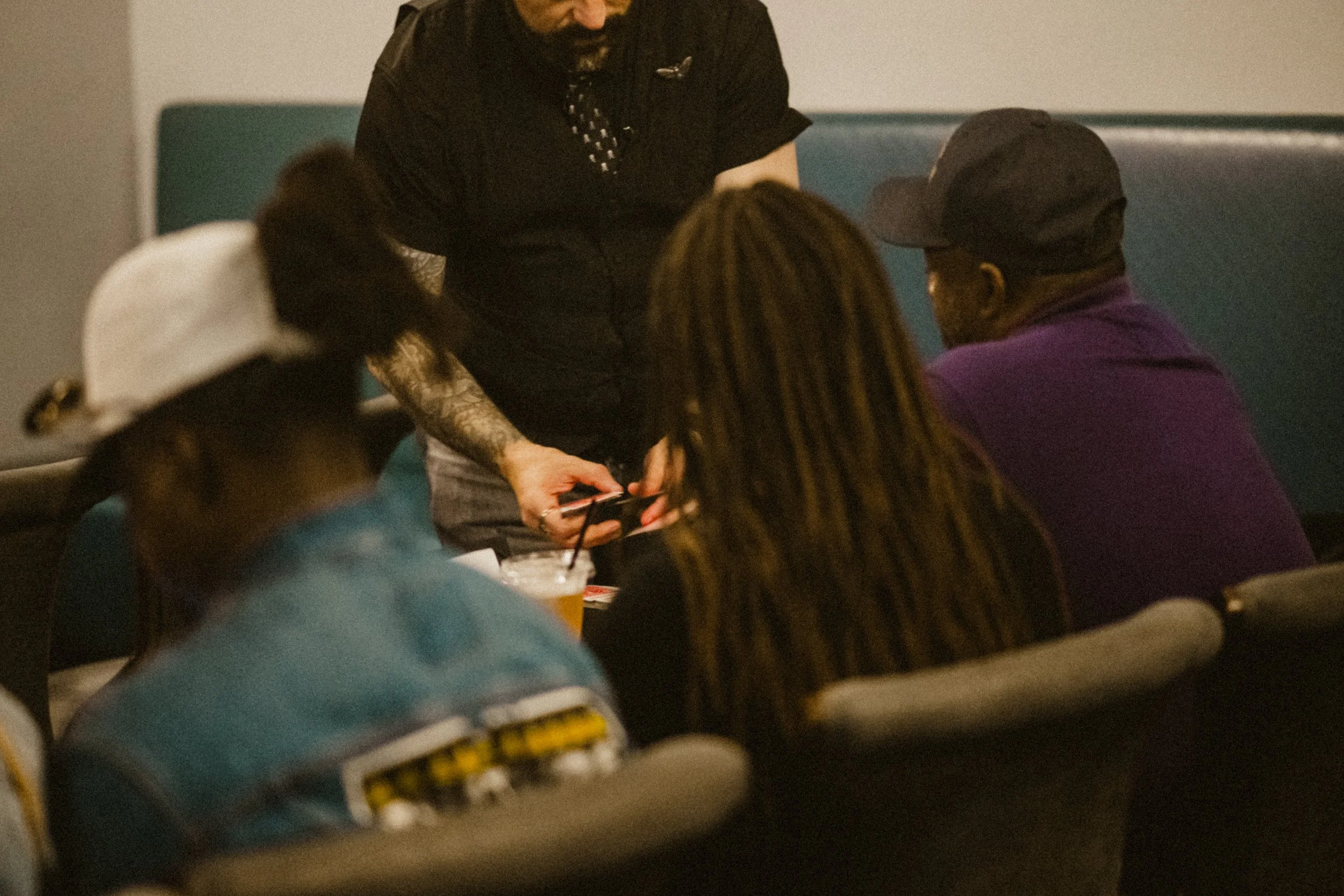 Group of people sitting together in a restaurant or cafe, with a magician standing and showing a card trick to the group.
