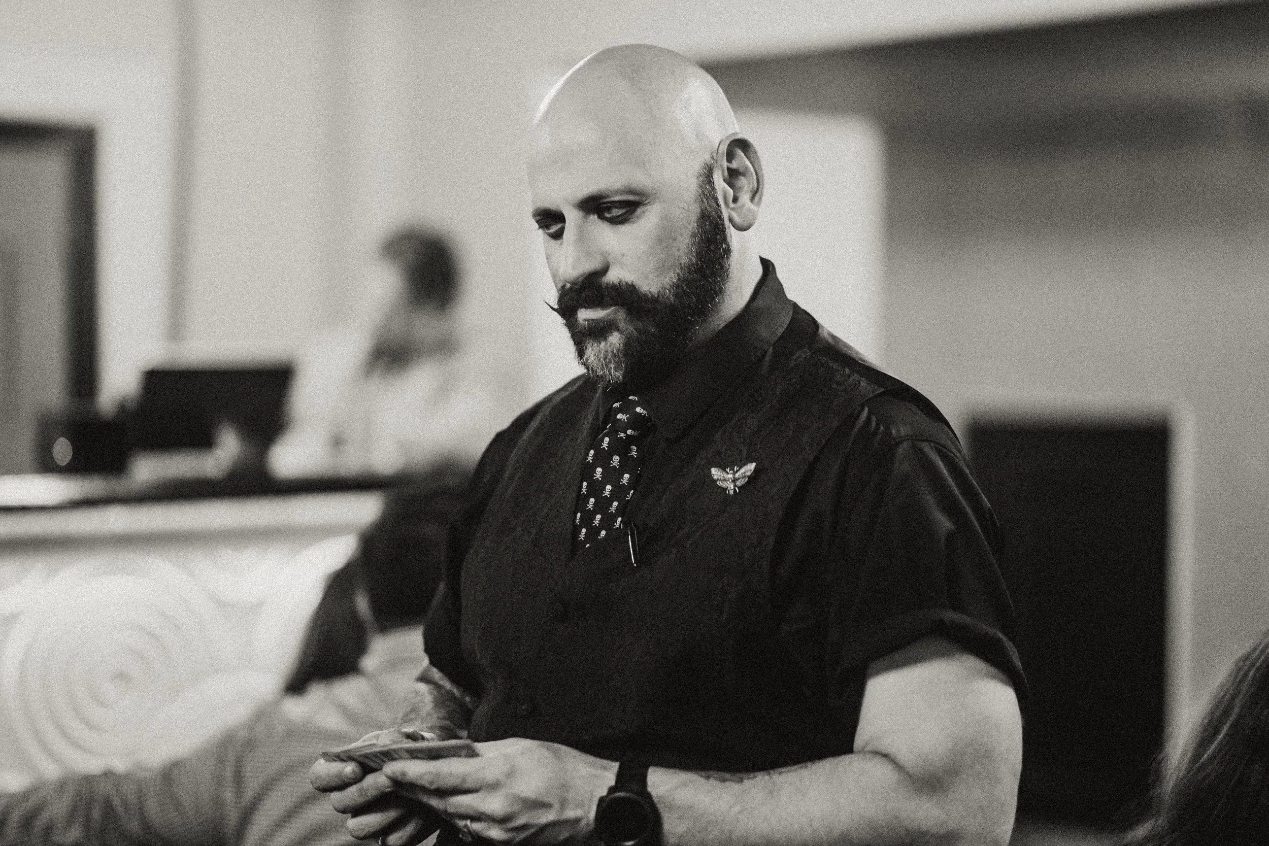 A bald man with a beard and mustache, wearing a dark shirt, vest, and tie with a skull pattern, is looking down at his phone in an indoor setting.