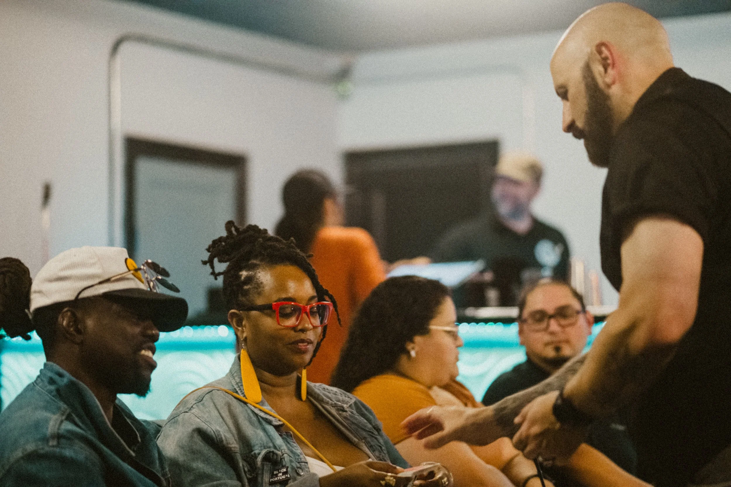 A magician with a shaved head and beard interacting with a group of diverse people sitting down in a social setting, possibly a bar or cafe, with others in the background.