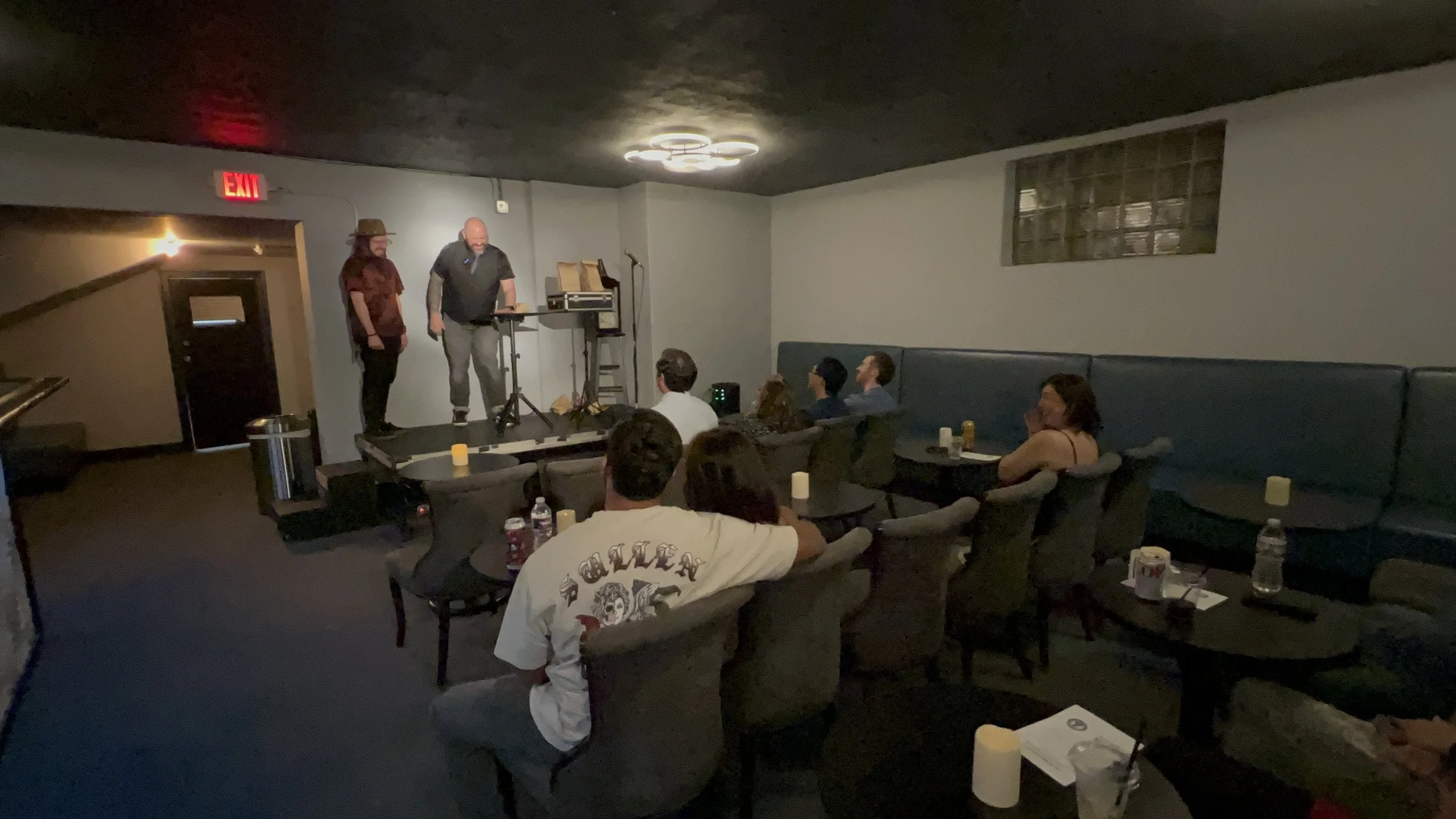 A small comedy or music performance in a dimly lit room with an audience seated at tables, watching two performers on stage. The performers are standing near a microphone and sound equipment, with some audience members looking towards the stage.