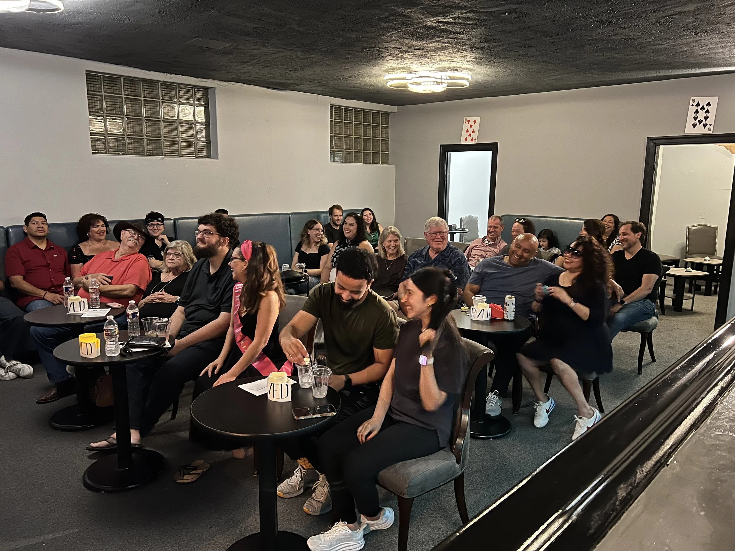 Group of people seated and laughing in a room with playing cards on the wall, some with drinks on the tables, indicating a casual social gathering.