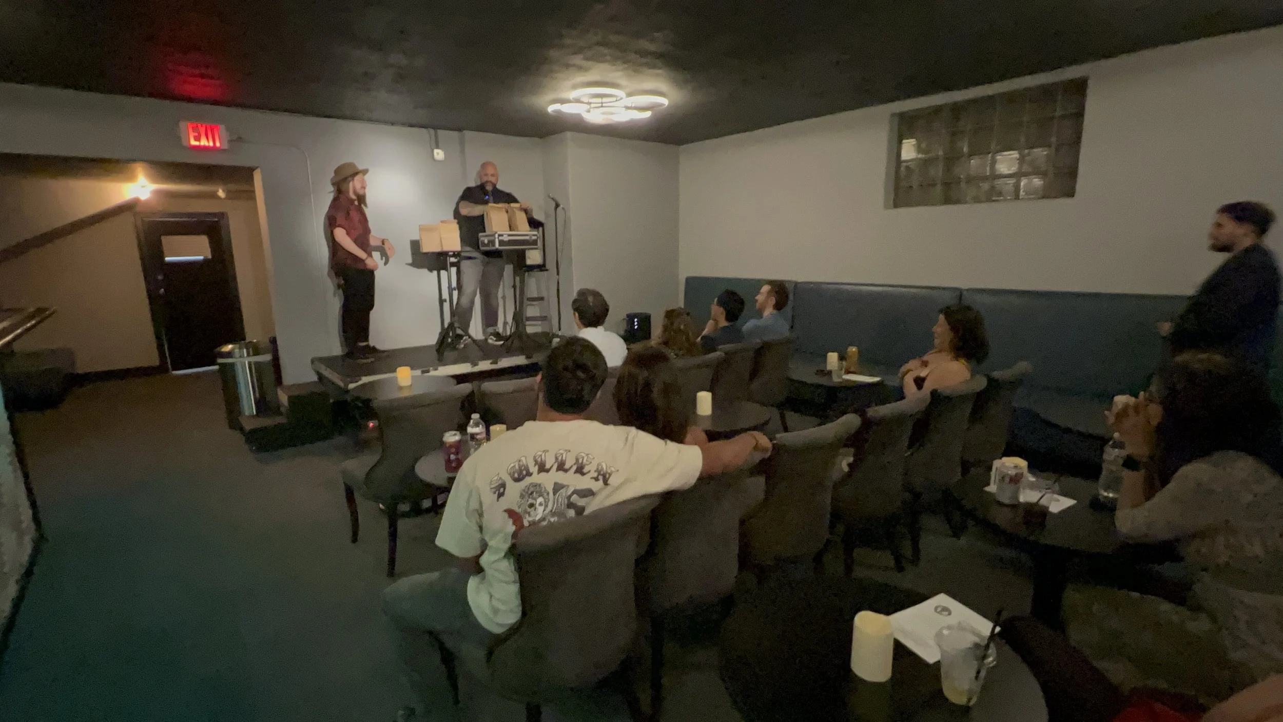 A comedy show in a small, dimly lit venue featuring two performers on stage, one holding a microphone and a box, with an audience seated at tables watching.