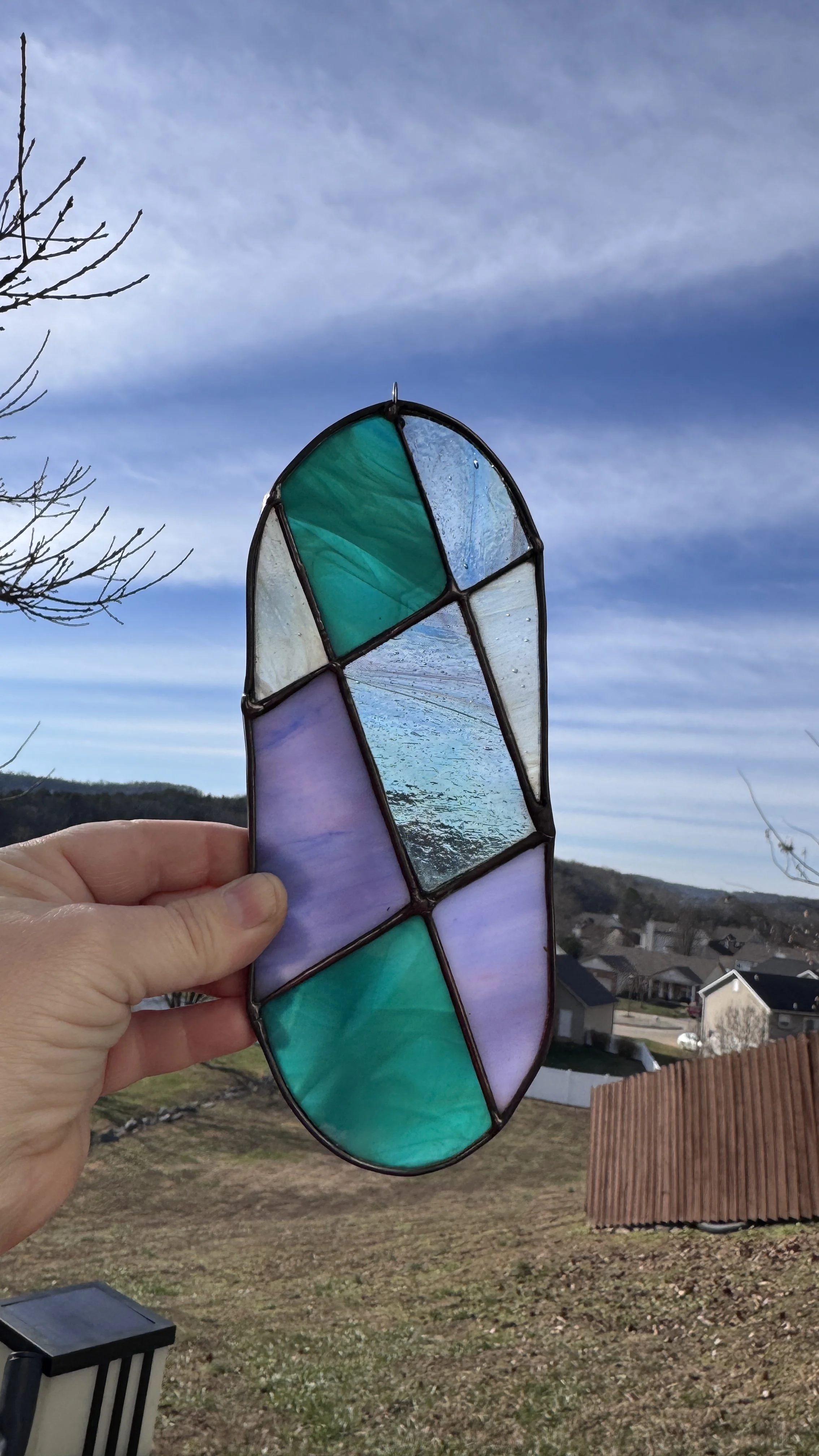 Hand holding a stained glass piece with geometric shapes in various shades of blue, green, and purple against a sky background with clouds and a suburban neighborhood.