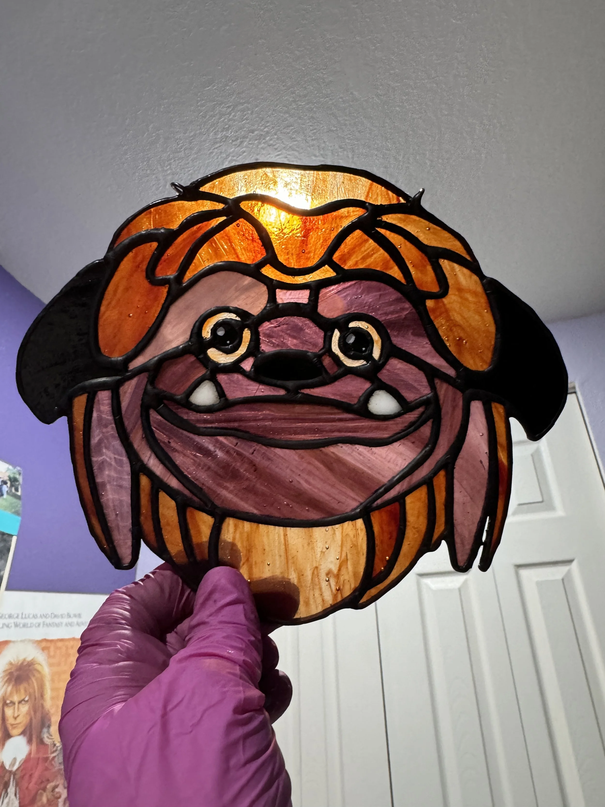 Stained glass lamp in the shape of a dog's face, held by a person wearing pink gloves, with a purple wall and white closet doors in the background.
