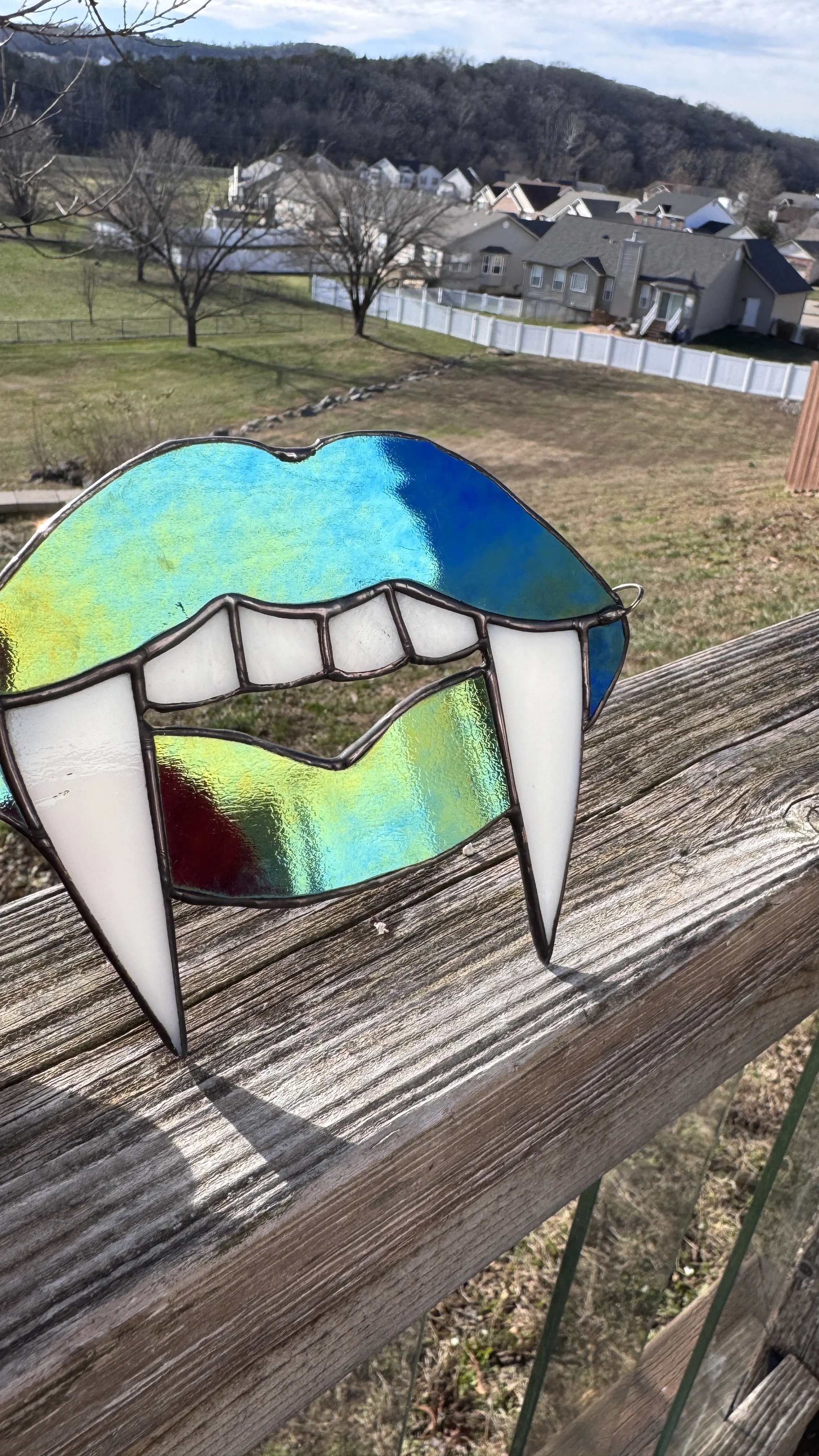 Close-up of a stained glass design resembling an open mouth with teeth, placed on a wooden railing outdoors with houses and a hillside in the background.