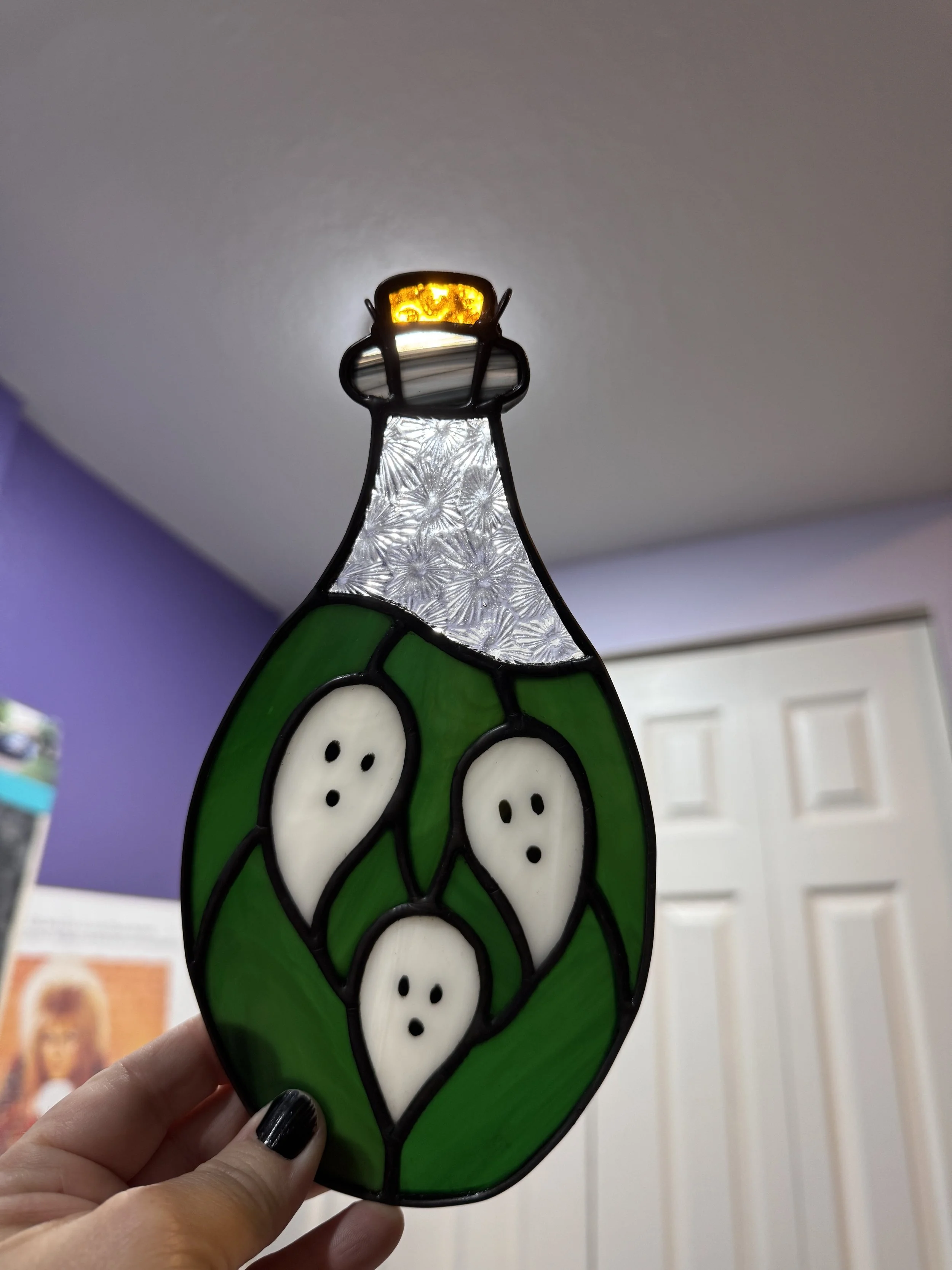 Close-up of a stained glass potion depicting ghostly figures with white faces, black eyes, and black mouths, set against a green background with a textured glass top, illuminated from within.
