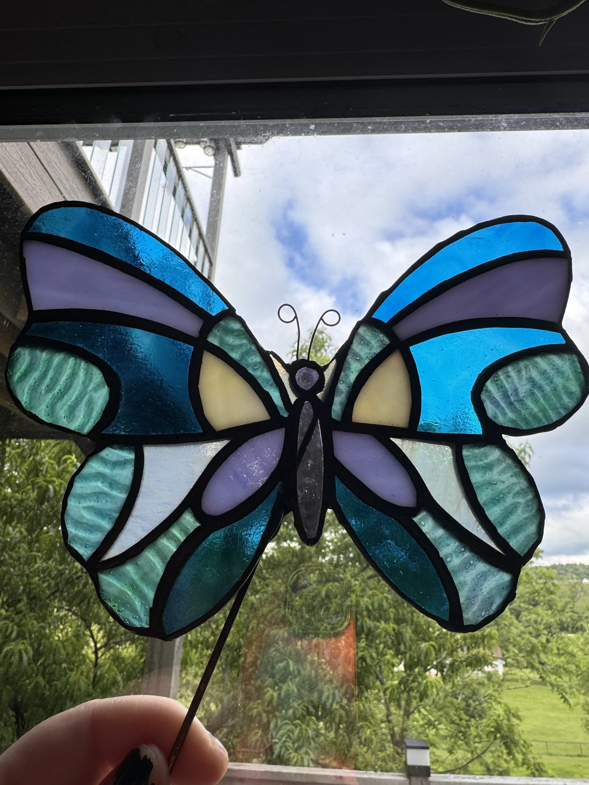 Hand holding a stained glass butterfly decoration in front of a window with trees and a cloudy sky outside.