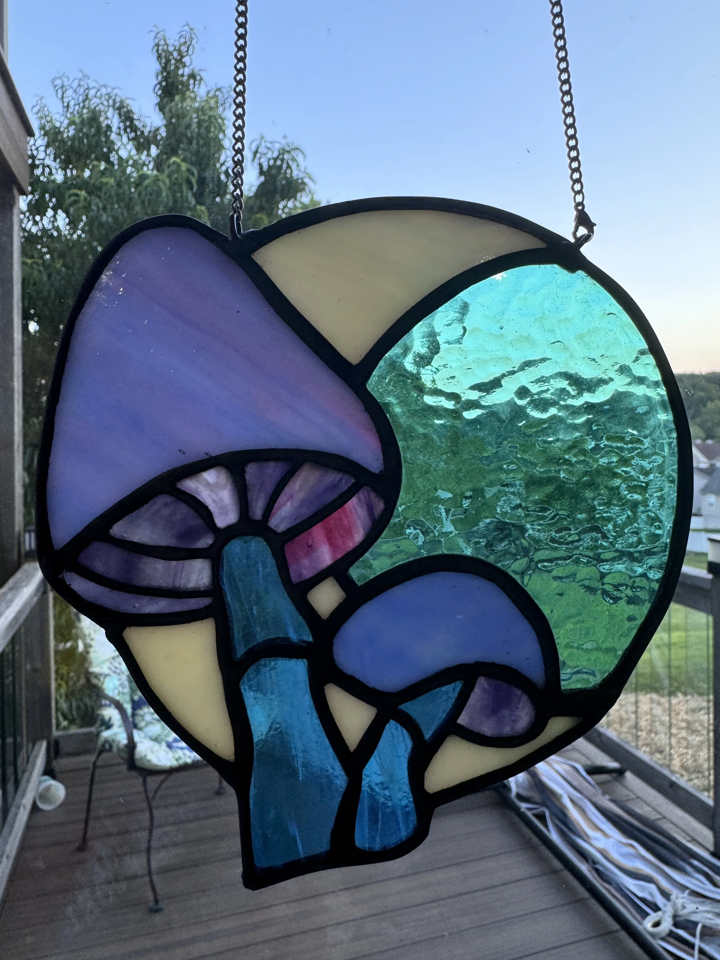 Colorful stained glass wind chime featuring a mushroom, a tree, and abstract shapes, hanging outdoors against a blue sky with trees in the background.