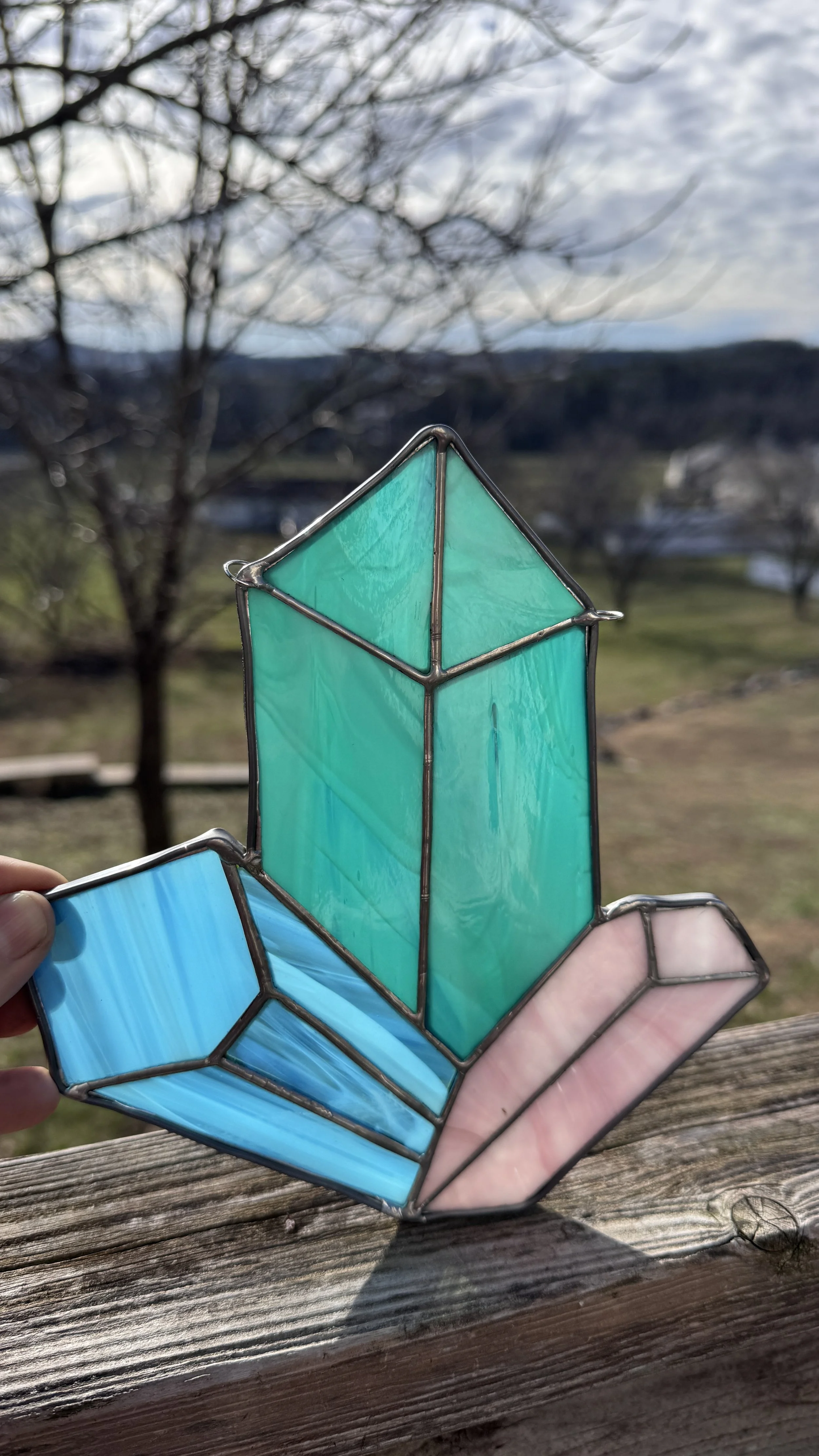 Colorful stained glass stained glass sculpture with blue, pink, and turquoise pieces, held up outdoors against a background of a blurred landscape with trees and sky.