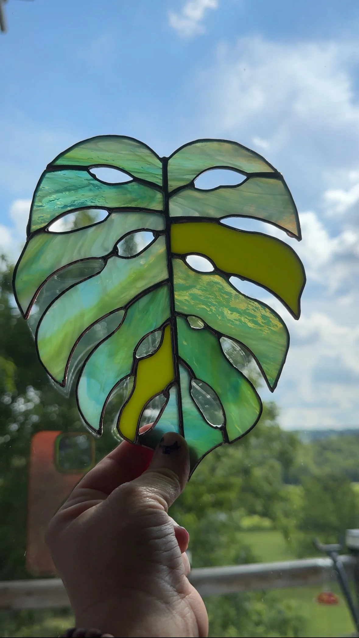 A stained glass lettuce leaf decoration being held up against a blue sky with clouds, with trees and a railing in the background.