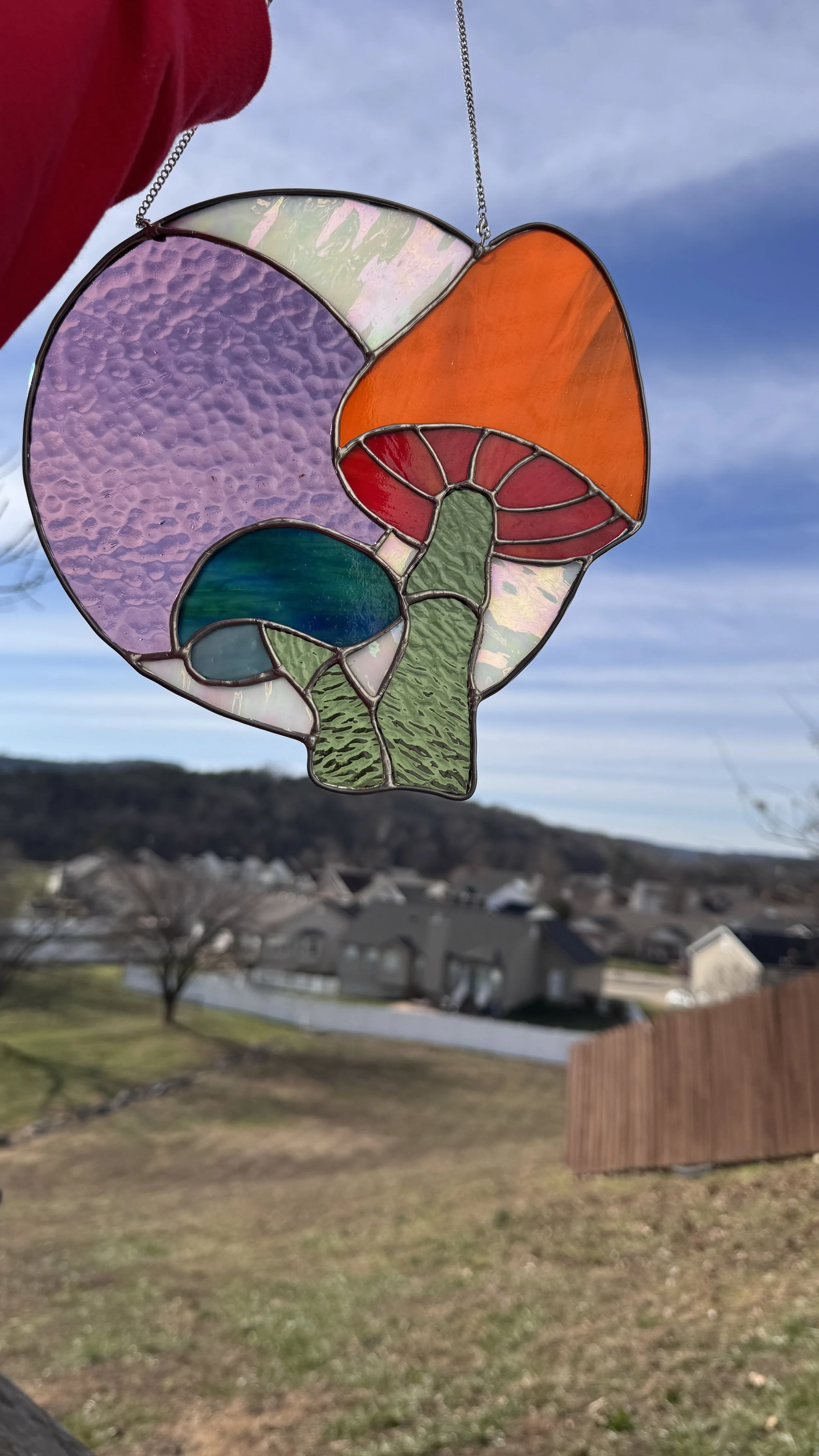 Colorful stained glass sun catcher with mushrooms design held against a clear blue sky with a suburban backyard in the background.