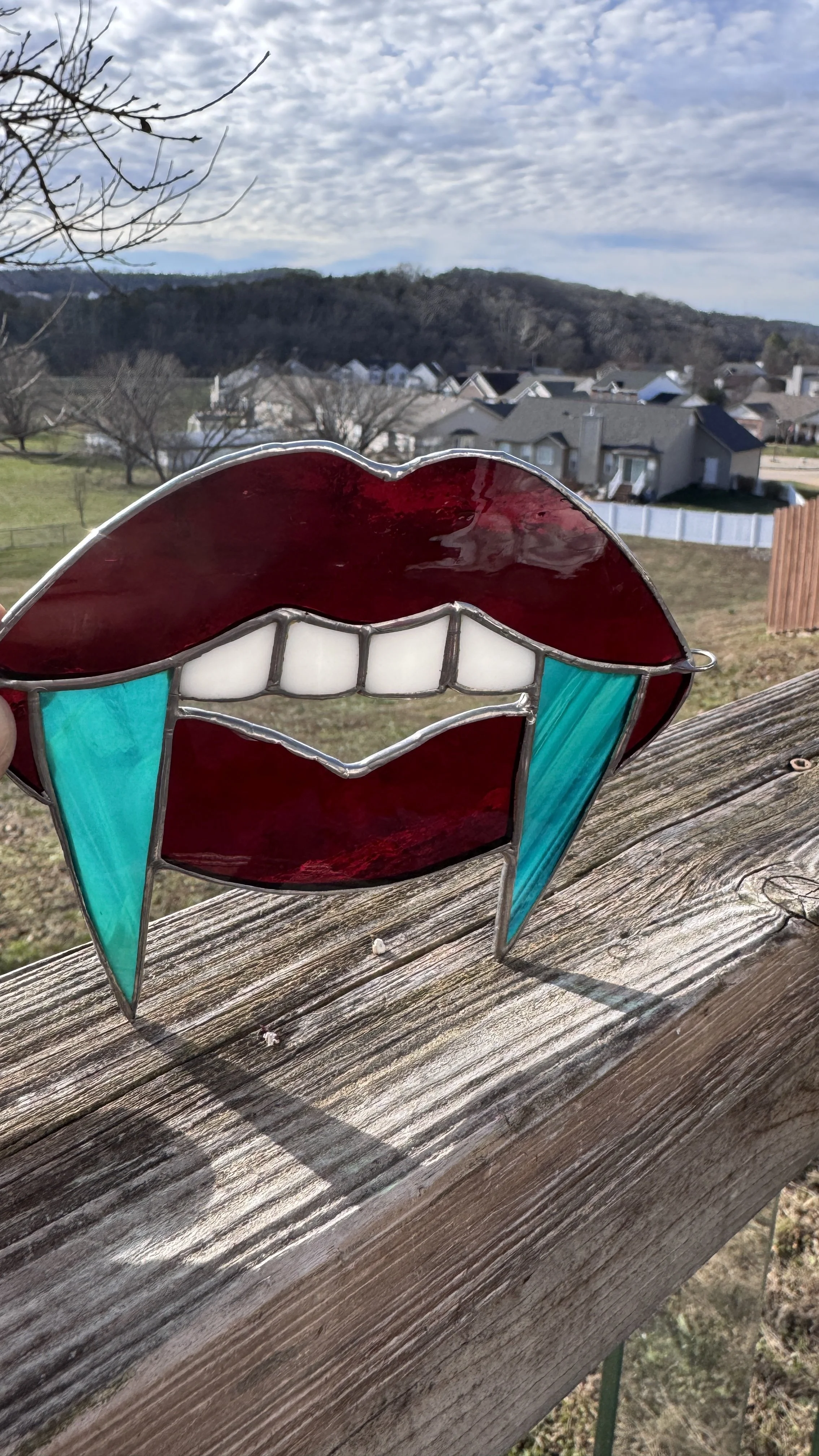 A stained glass piece in the shape of lips with teeth, featuring red, white, and teal glass, placed on a wooden surface outdoors, with a view of a neighborhood and mountains in the background.