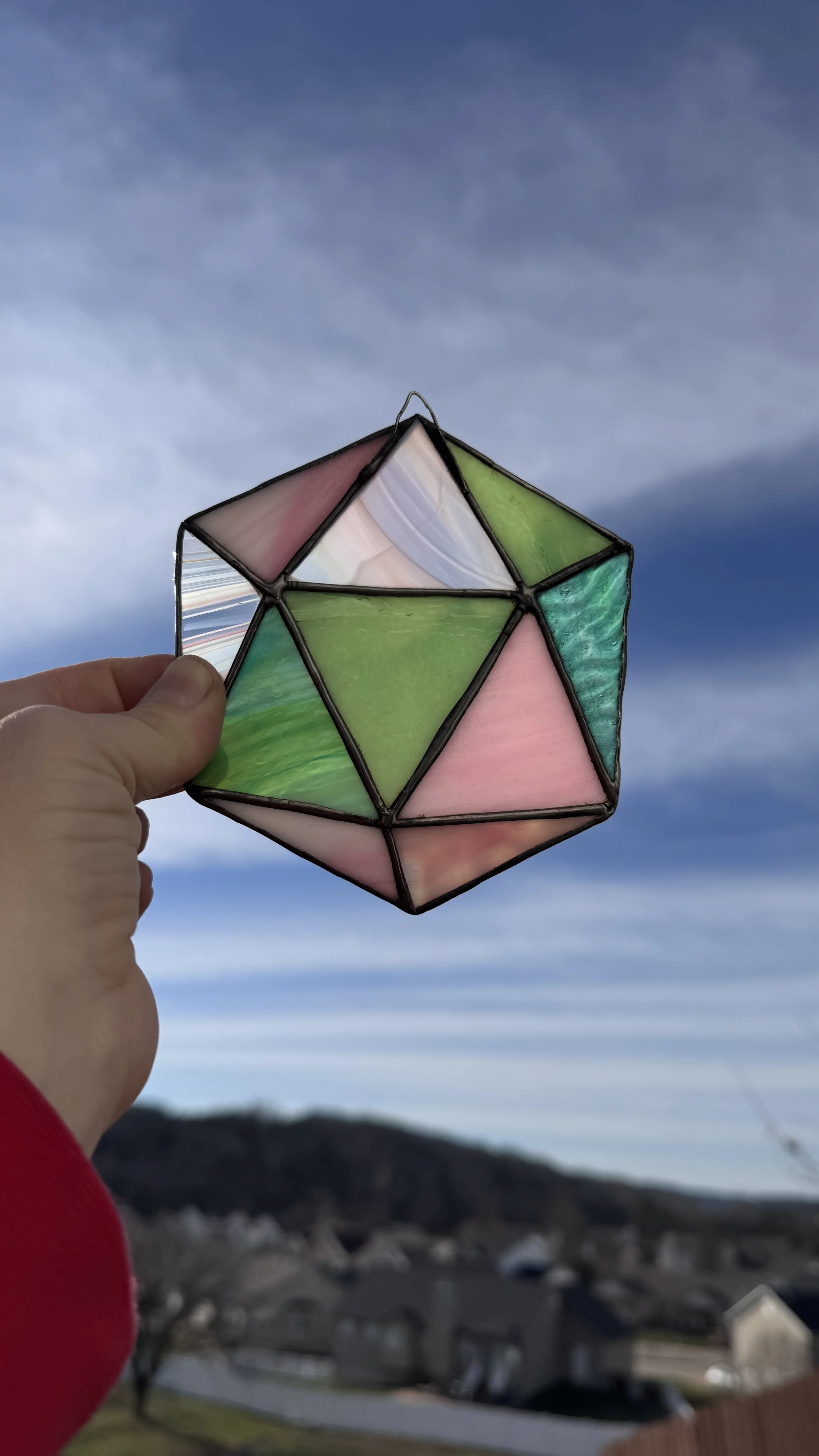 Person holding a stained glass geometric pendant against the sky, with houses and a mountain in the background.