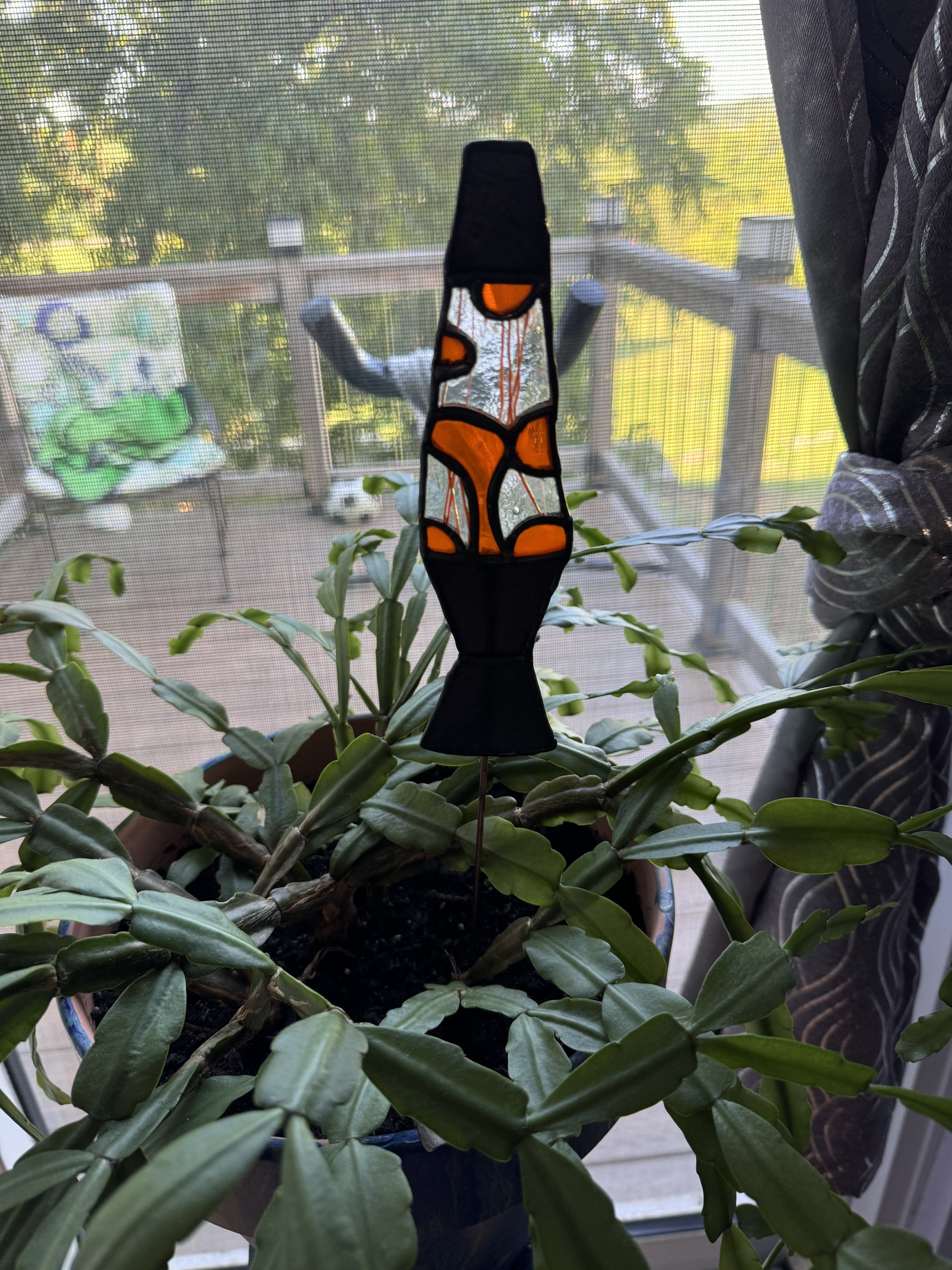 A potted plant with elongated dark green leaves on a balcony with a mesh screen and a stained glass lava lamp in front of it.