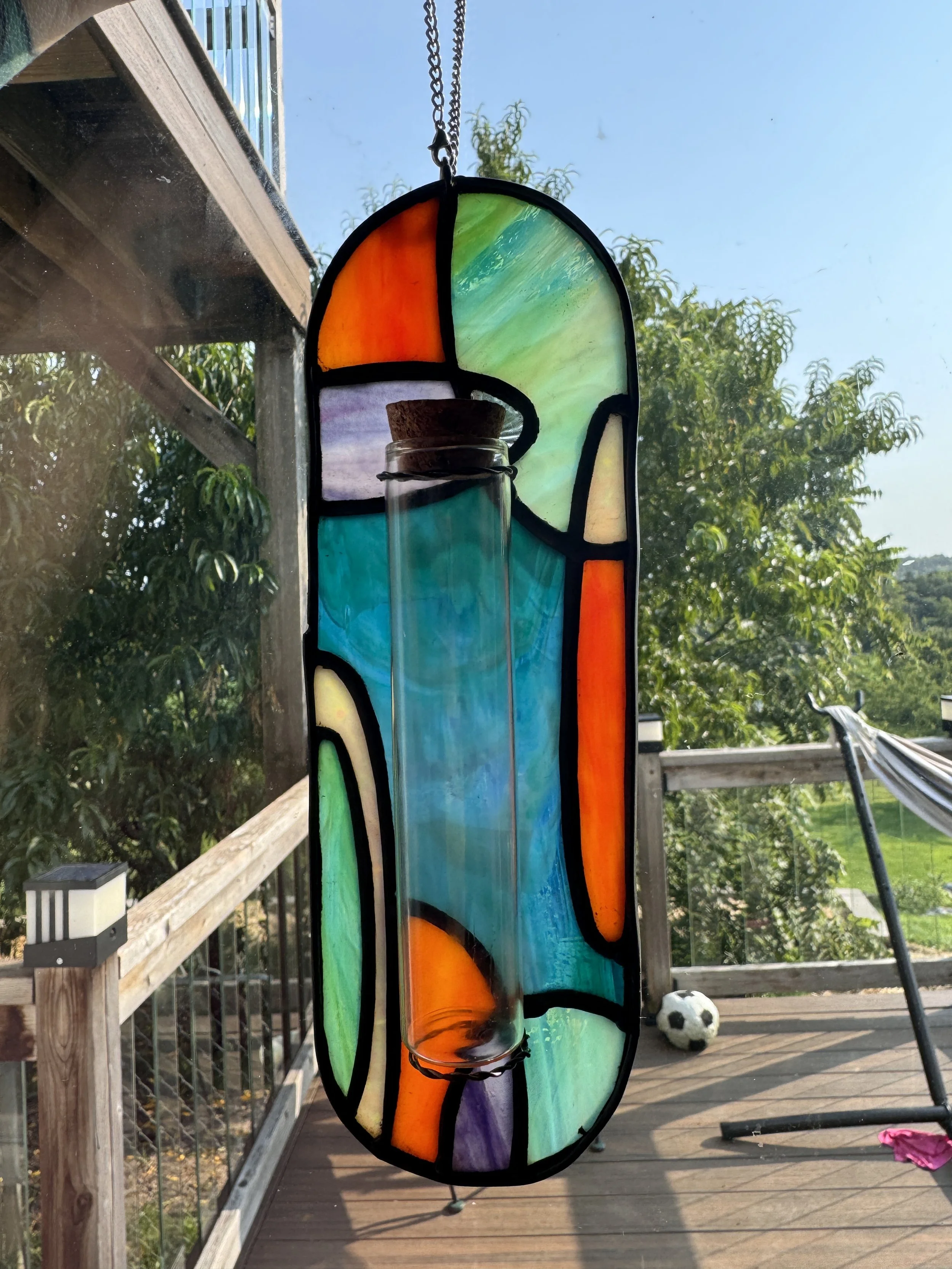 Colorful stained glass hanging ornament with a test tube attached, outdoors on a wooden deck with trees, a soccer ball, and a hammock visible in the background.