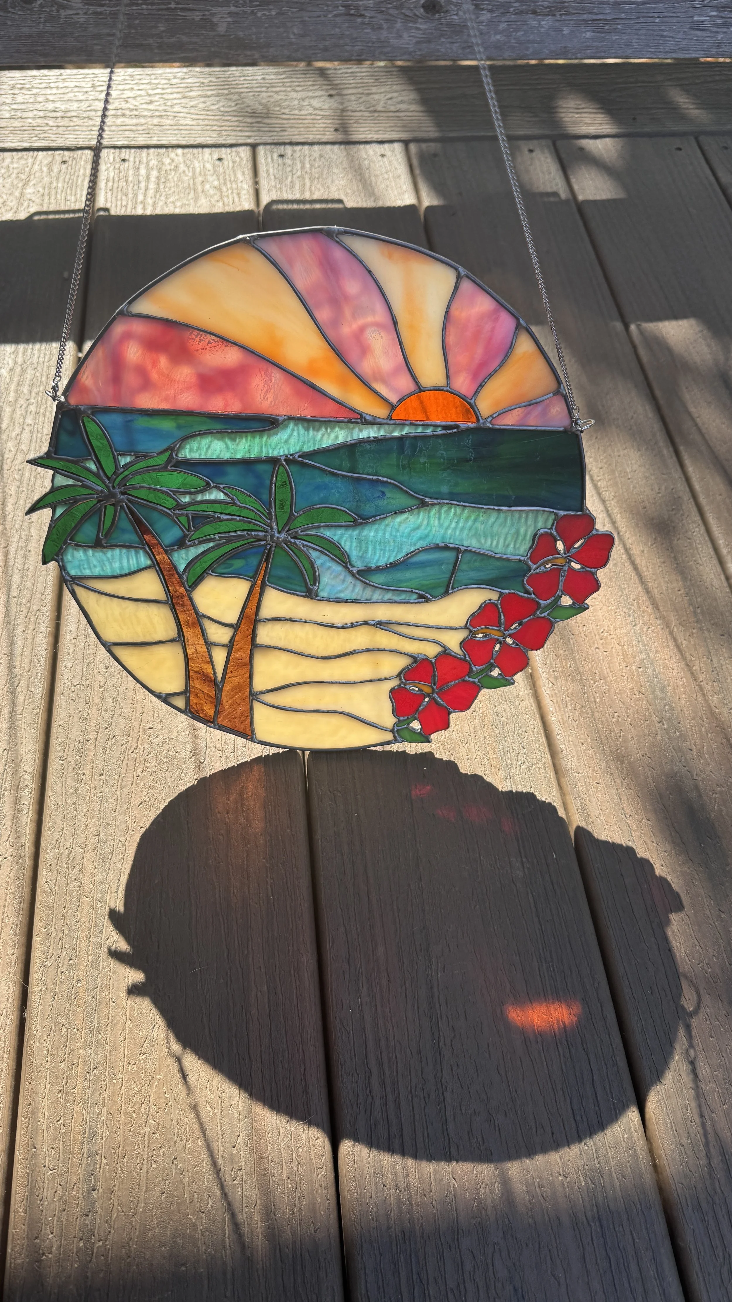 Colorful stained glass sun and ocean scene with palm trees and red flowers, hanging on a string with sunlight casting a shadow on the wooden surface.