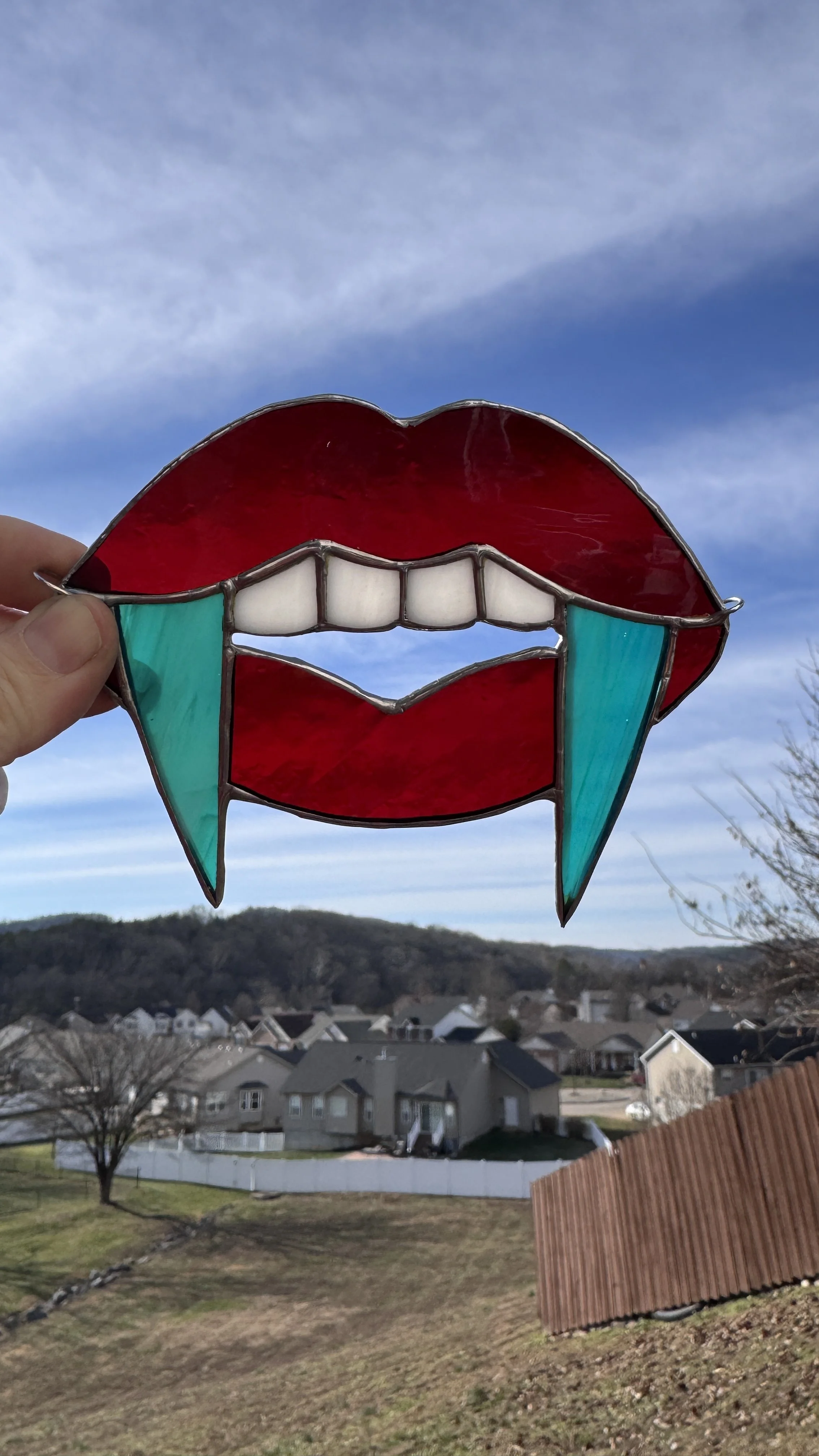 Colorful stained glass art piece depicting lips with teeth and tears, held up against a blue sky with houses and trees in the background.