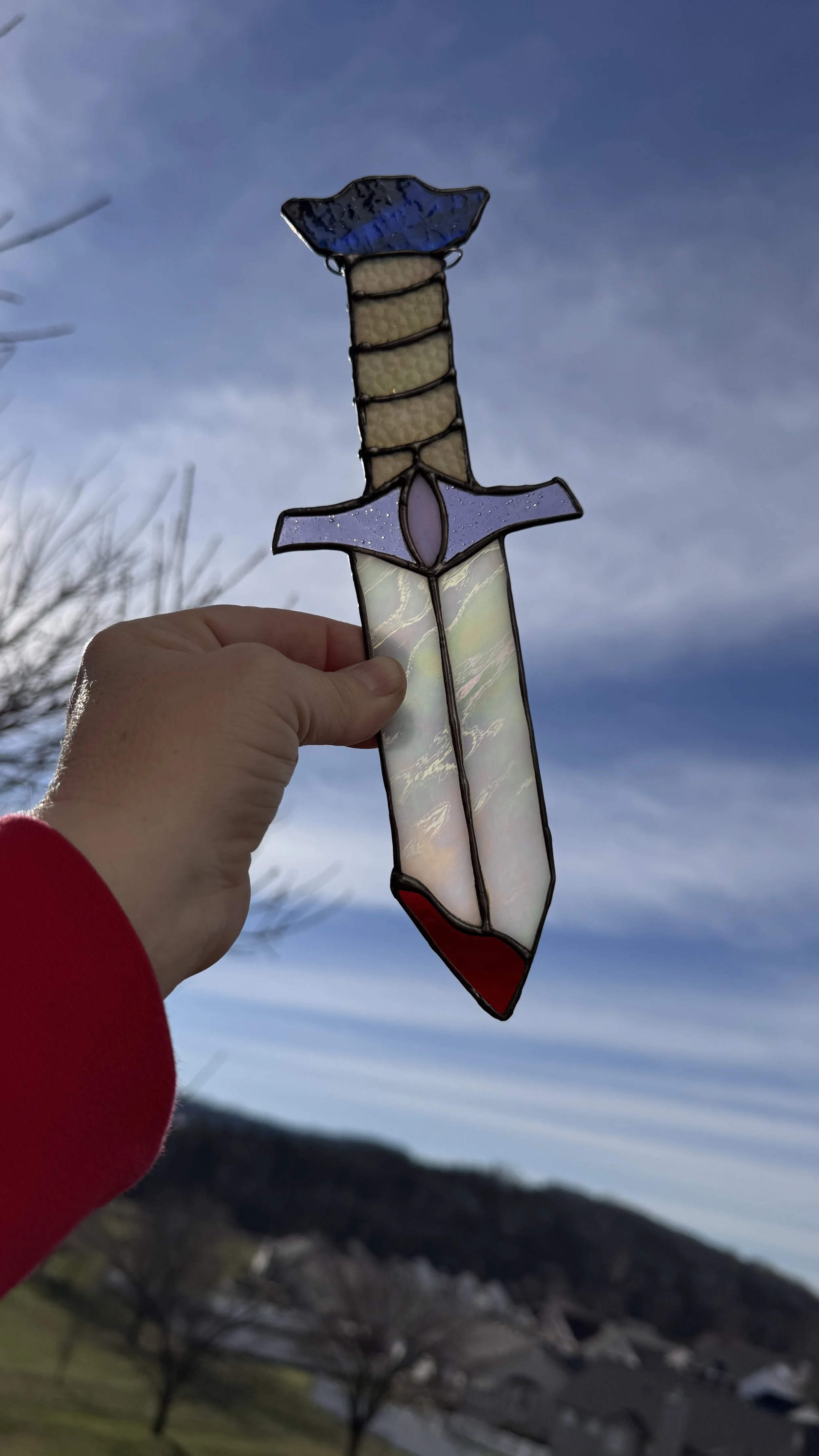 A stained glass sword held up against a cloudy sky, with mountains and trees in the background.
