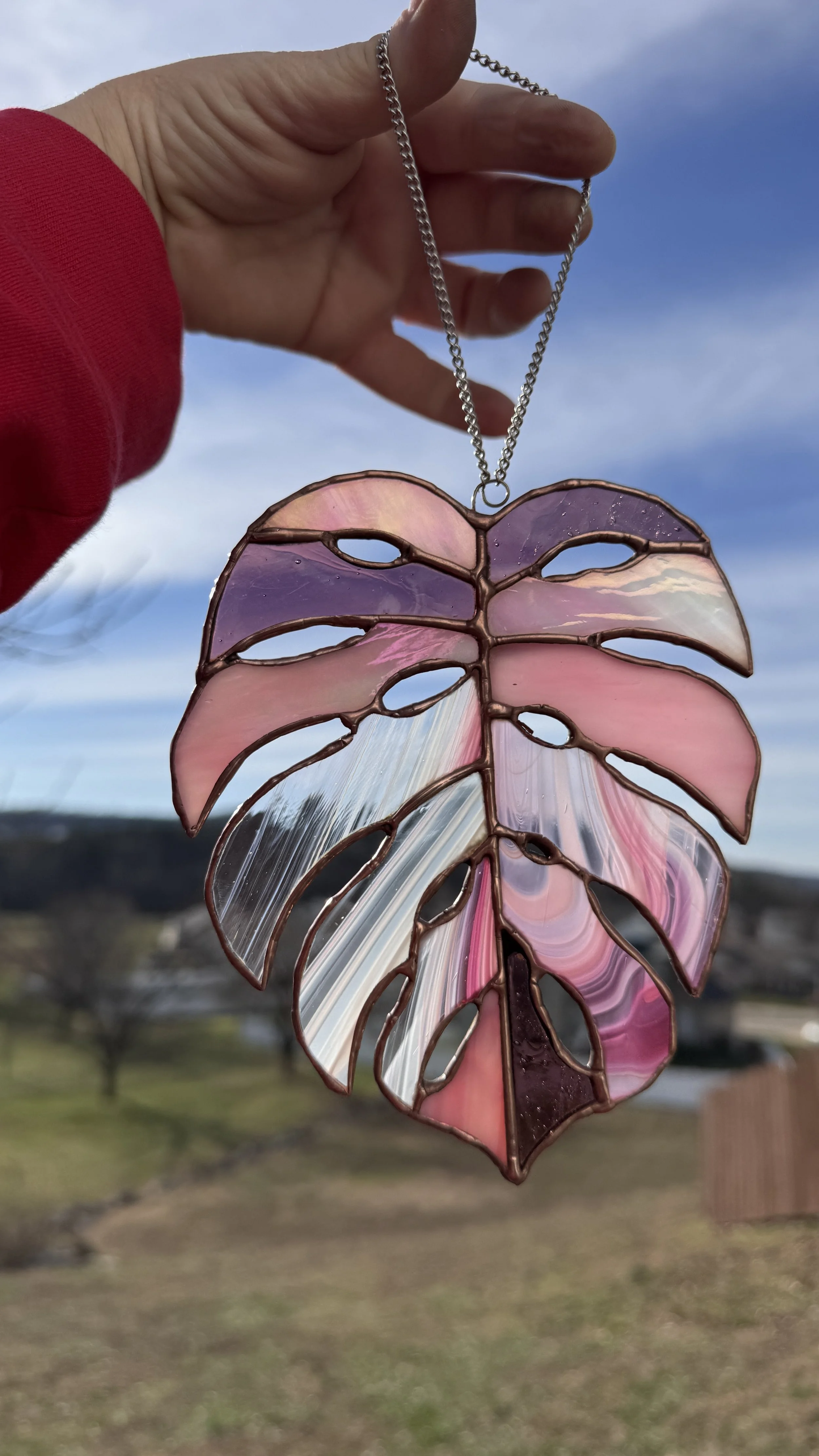 A stained glass pendant shaped like a large monstera leaf, with pink, purple, and white swirls, held up outdoors against a blue sky with wispy clouds.