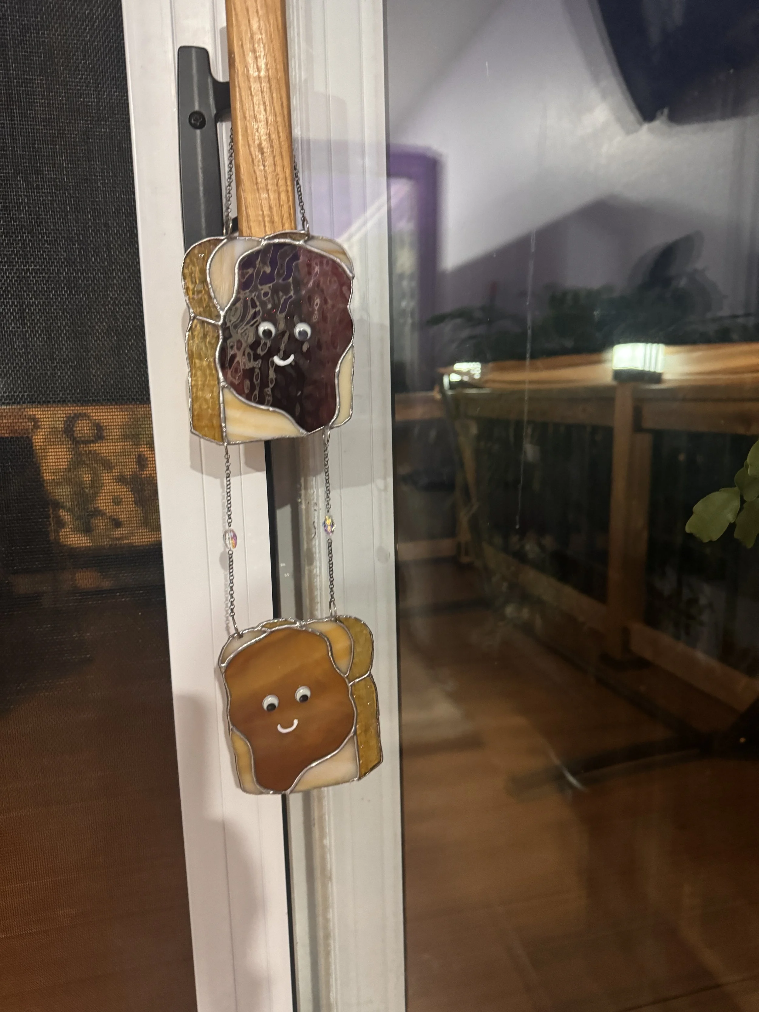 Stained glass window ornaments resembling peanut butter and jelly with googly eyes, smiling faces, and hanging chains, placed on a window.