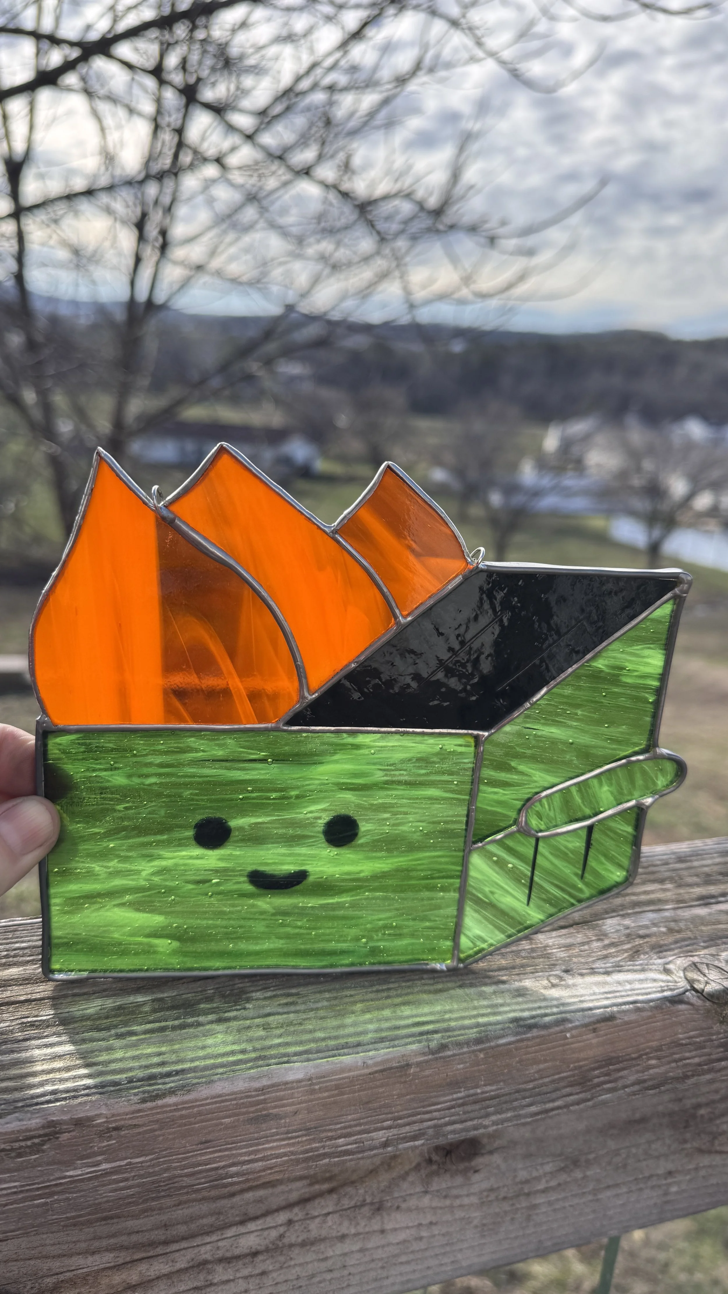 Colorful stained glass depiction of Baby Yoda from Star Wars, with orange ears, green face, and smiling expression, held outdoors on a wooden railing with a scenic landscape background.