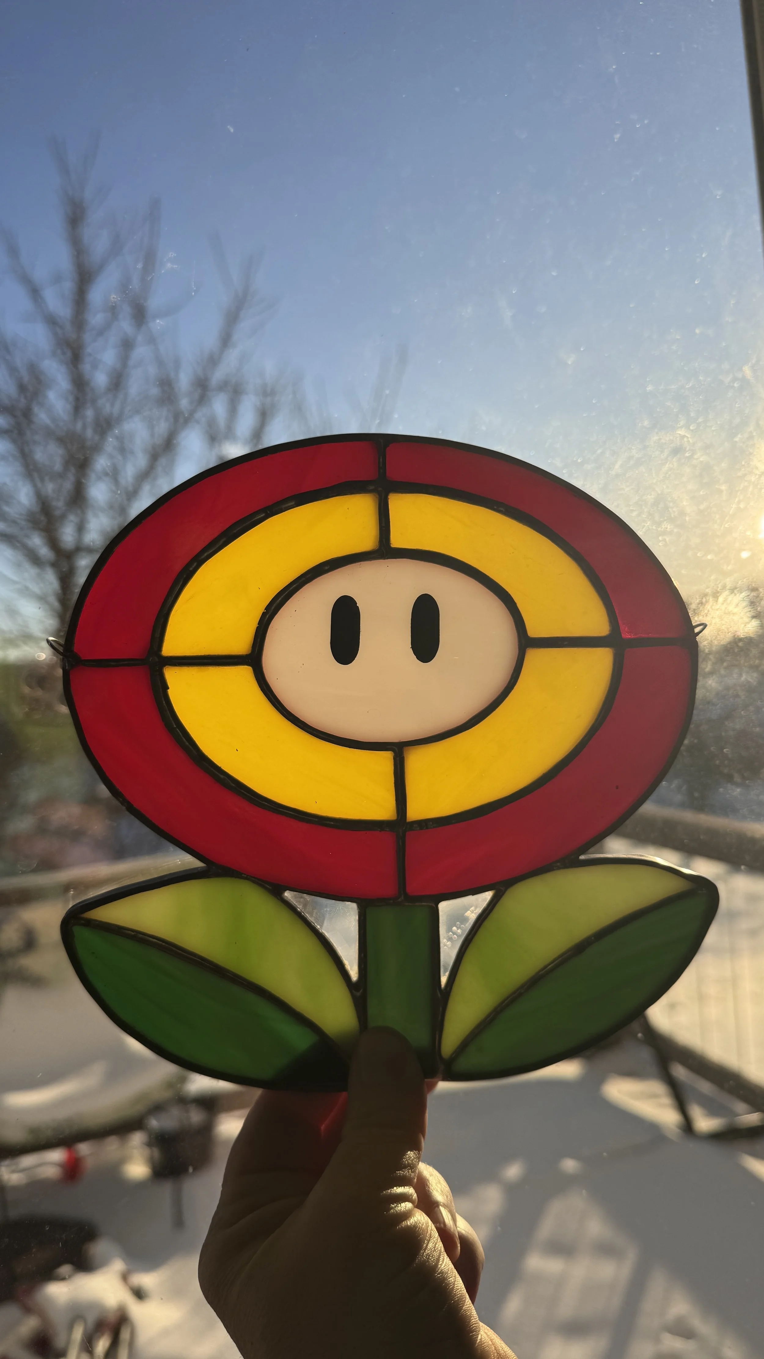 Colorful stained glass window in the shape of a flower with a pig face in the center, held up in front of a window with a snowy outdoor scene and a sunset or sunrise.