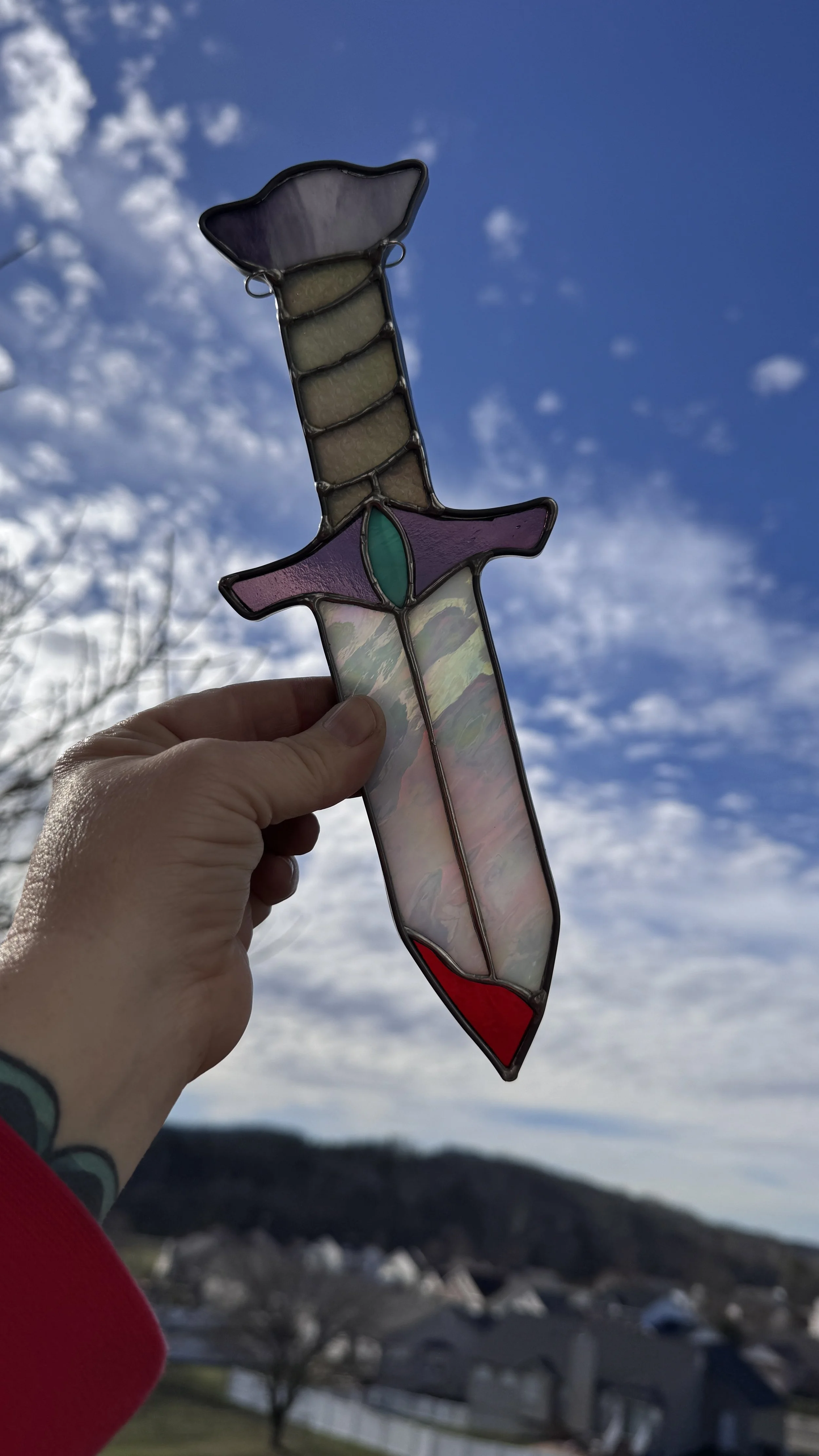 Colorful stained glass sword ornament being held up against the sky with some clouds and trees in the background.