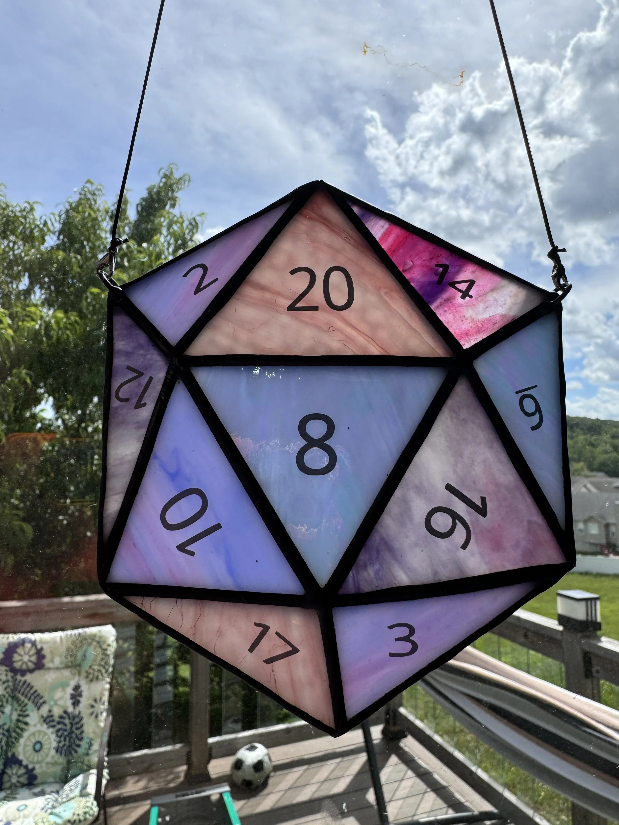 Colorful geometric hanging lantern with black number markings, floating against a cloudy blue sky, outdoor deck with chairs, cushions, and a small soccer ball.