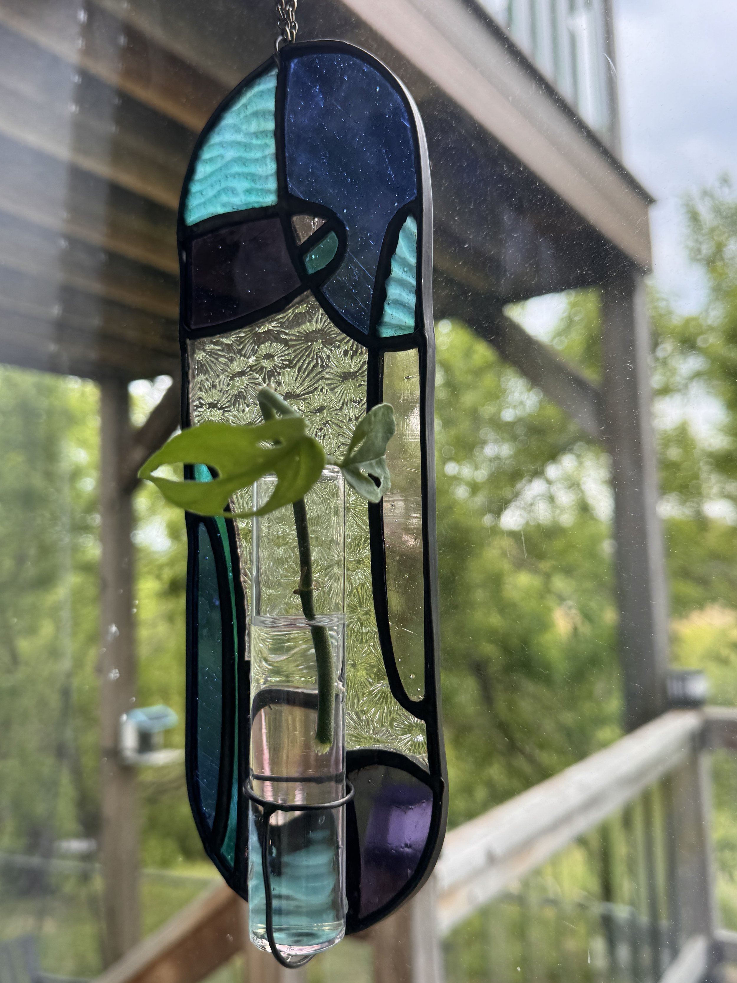 Stained glass plant propagation hanging near a window with trees outside.
