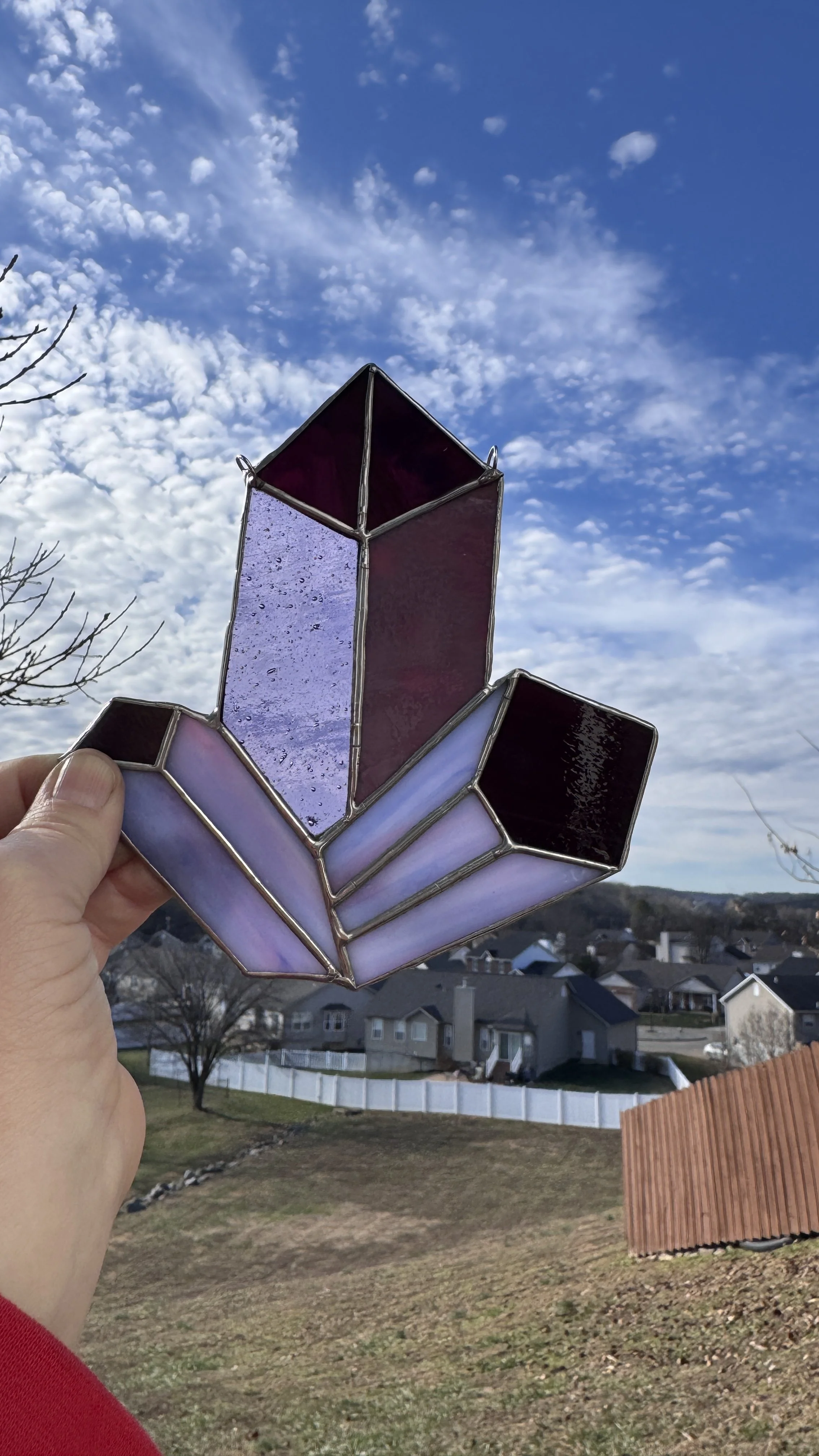 A hand holding a stained glass crystal art piece against a sky with scattered clouds and a residential neighborhood in the background.