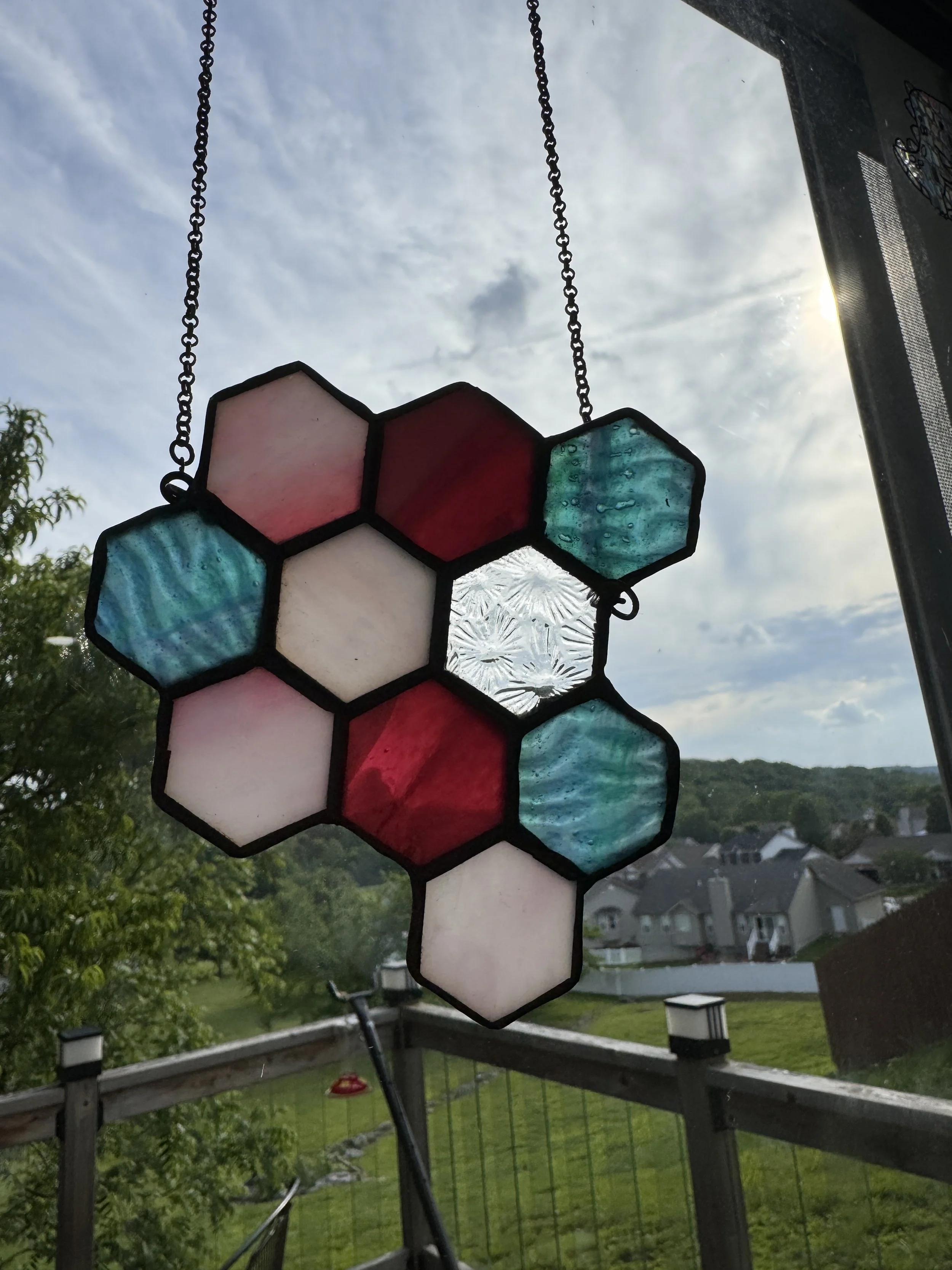 A stained glass decoration featuring hexagon shapes in pink, red, blue, and clear, hanging outdoors with a neighborhood and cloudy sky in the background.