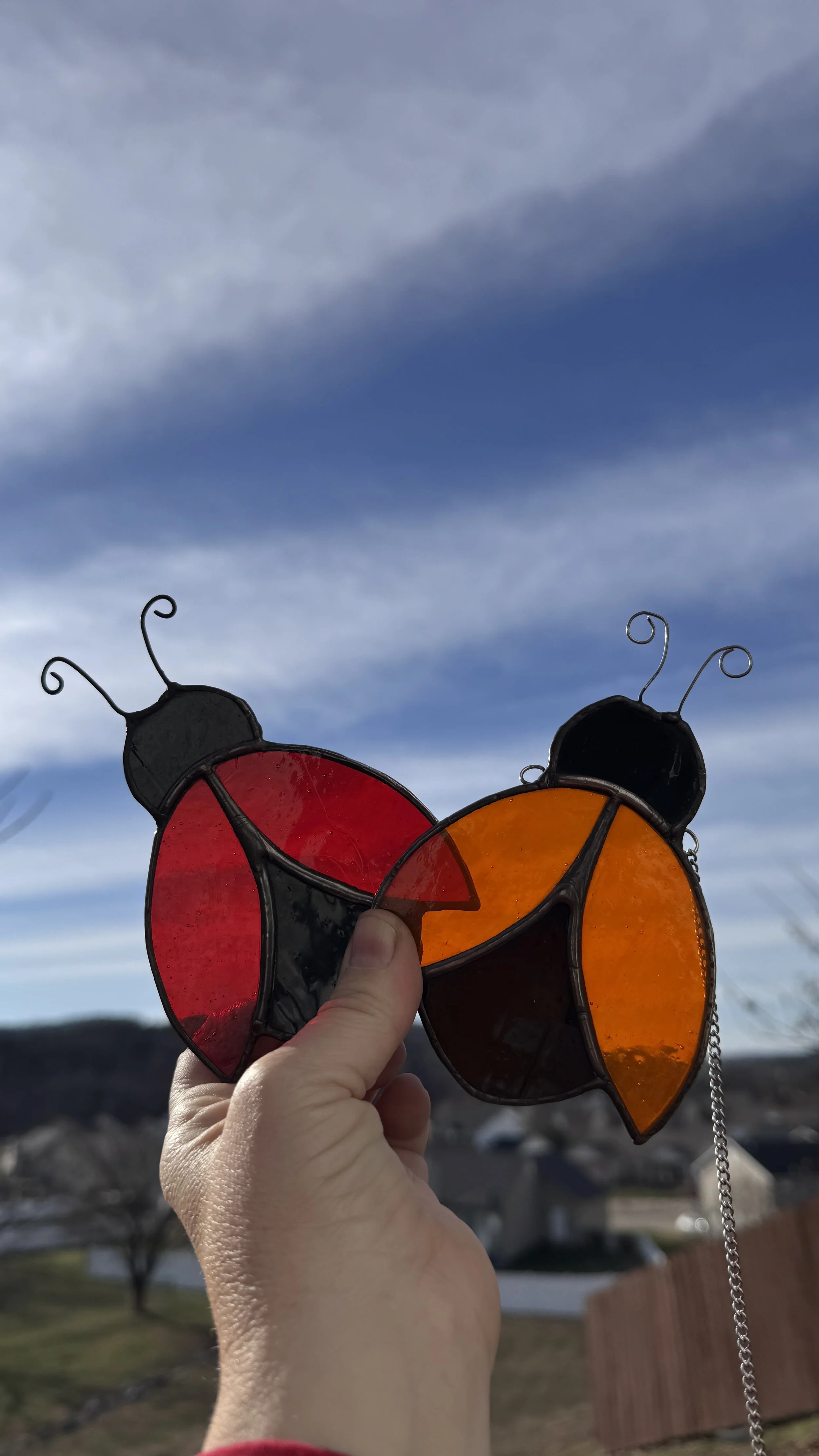 A hand holding stained glass  red and orange ladybugs against a cloudy sky and a suburban neighborhood in the background.