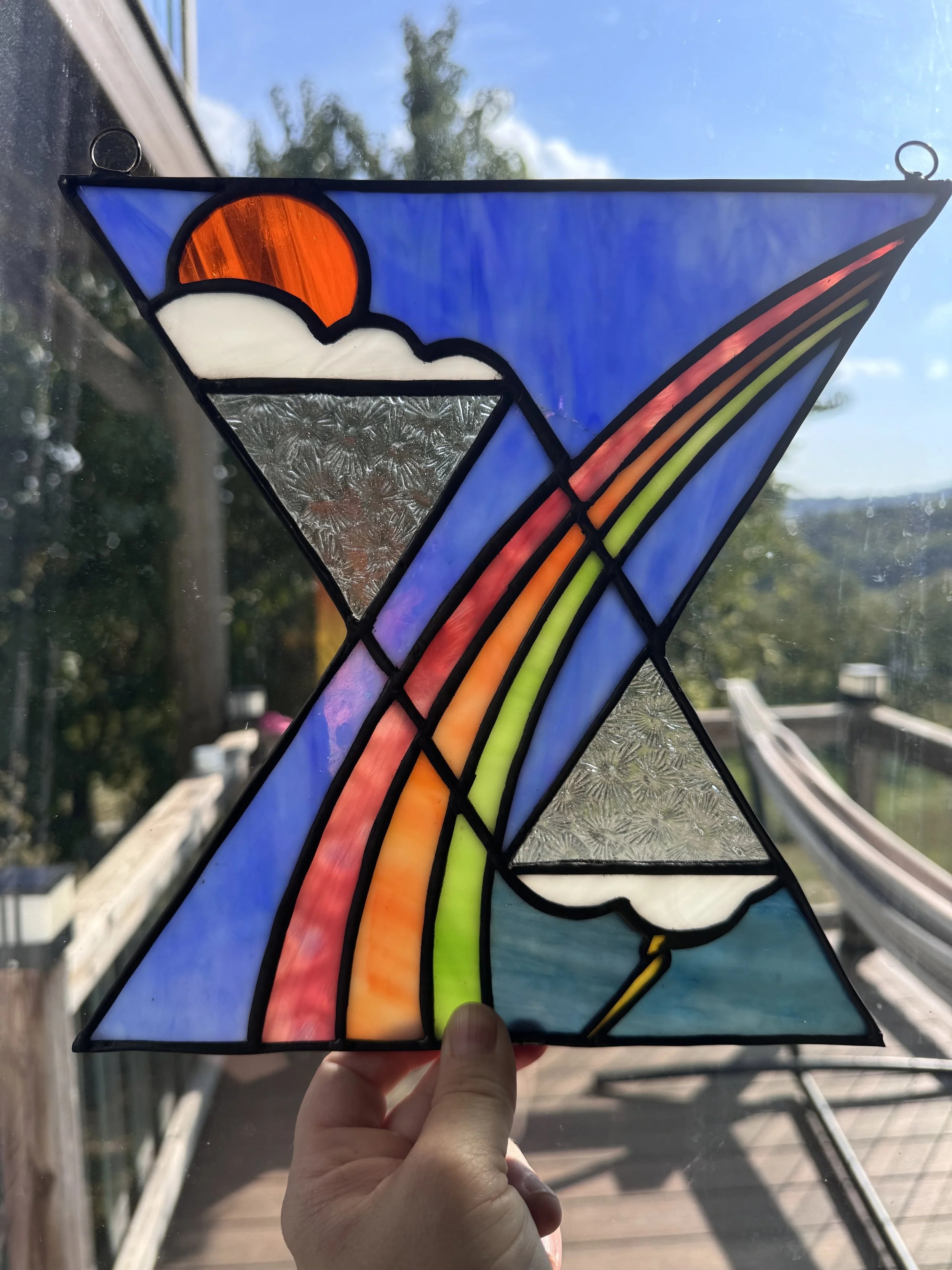 Stained glass artwork featuring a rainbow arc, clouds, blue sky, orange sun, and textured silver background, held up by a hand against the sky and deck background.