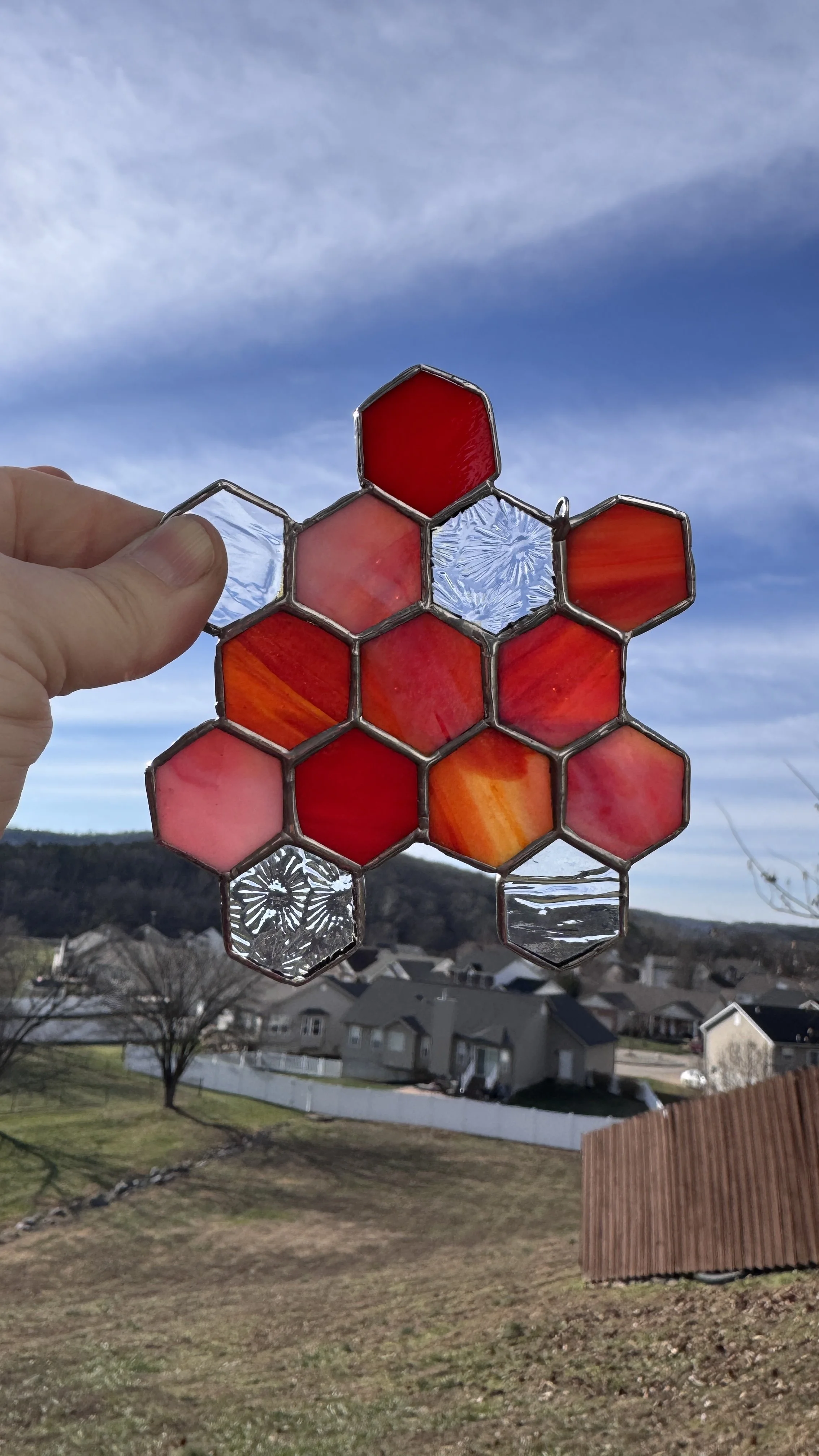 Hand holding a stained glass hexagon ornament against a clear blue sky with houses and trees in the background.