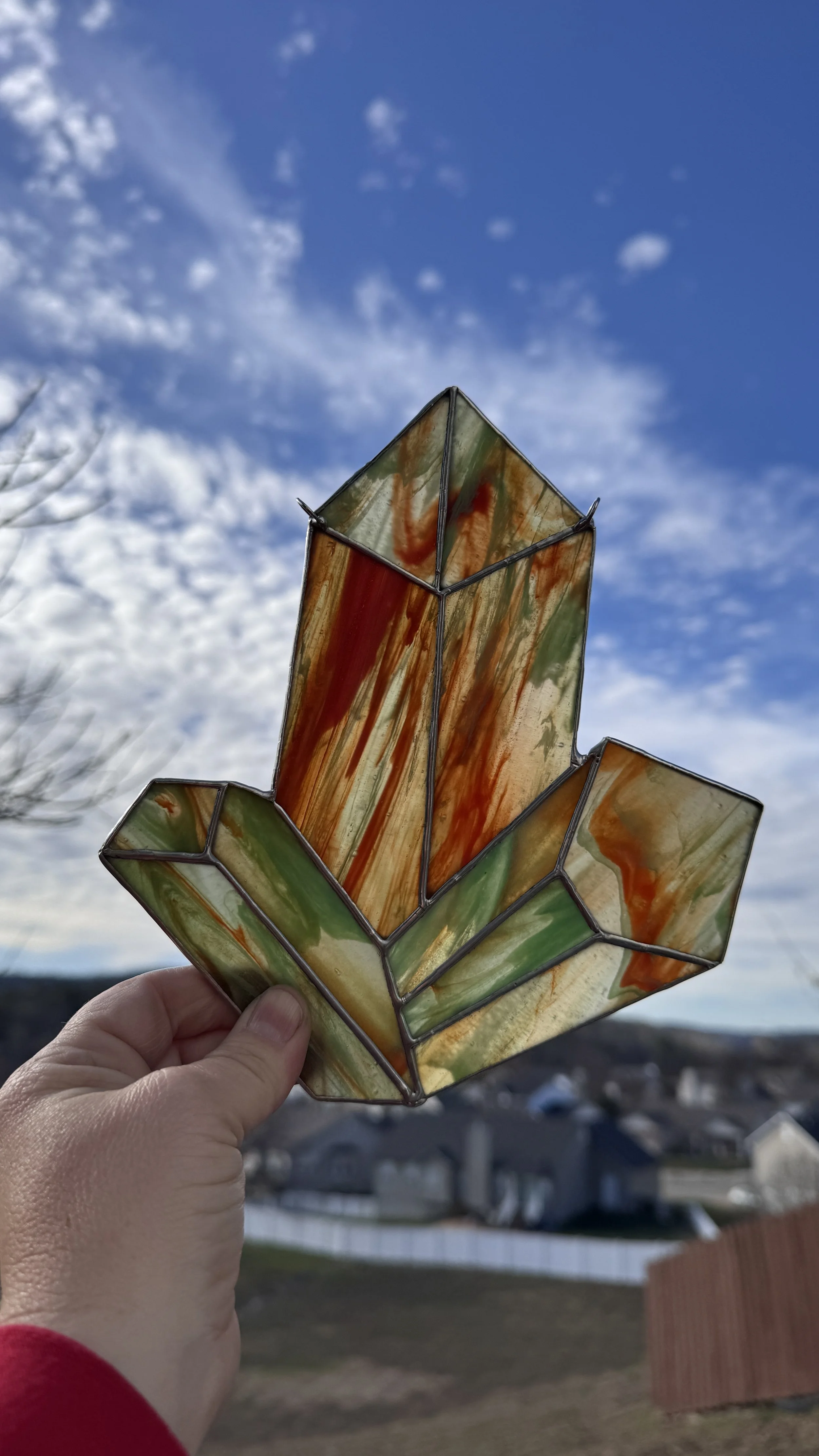Person holding a stained glass crystals that are orange and green t a blue sky with clouds, with houses and a yard in the background.