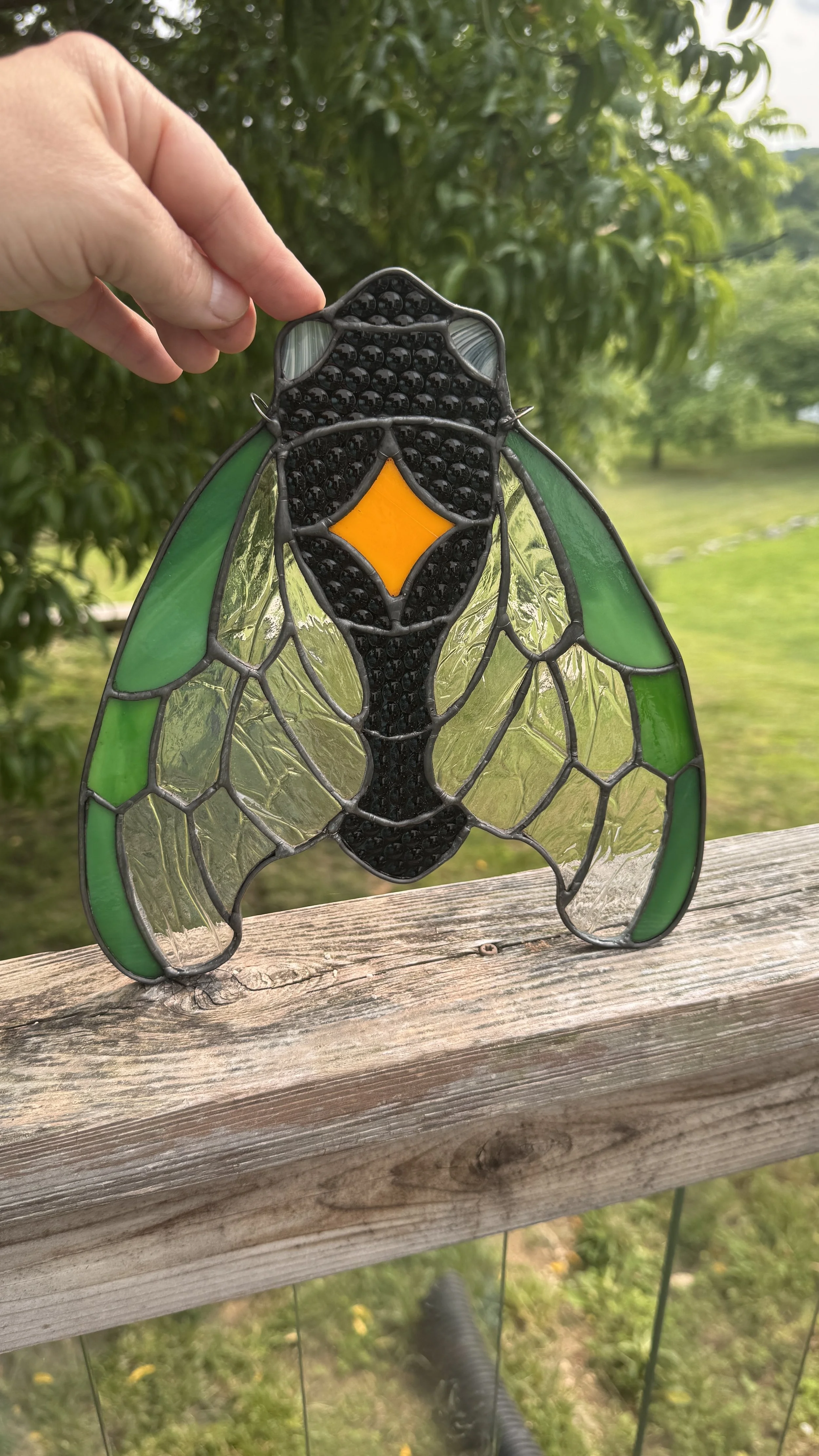 A stained glass piece resembling a cicada, held by a hand outdoors with greenery in the background.